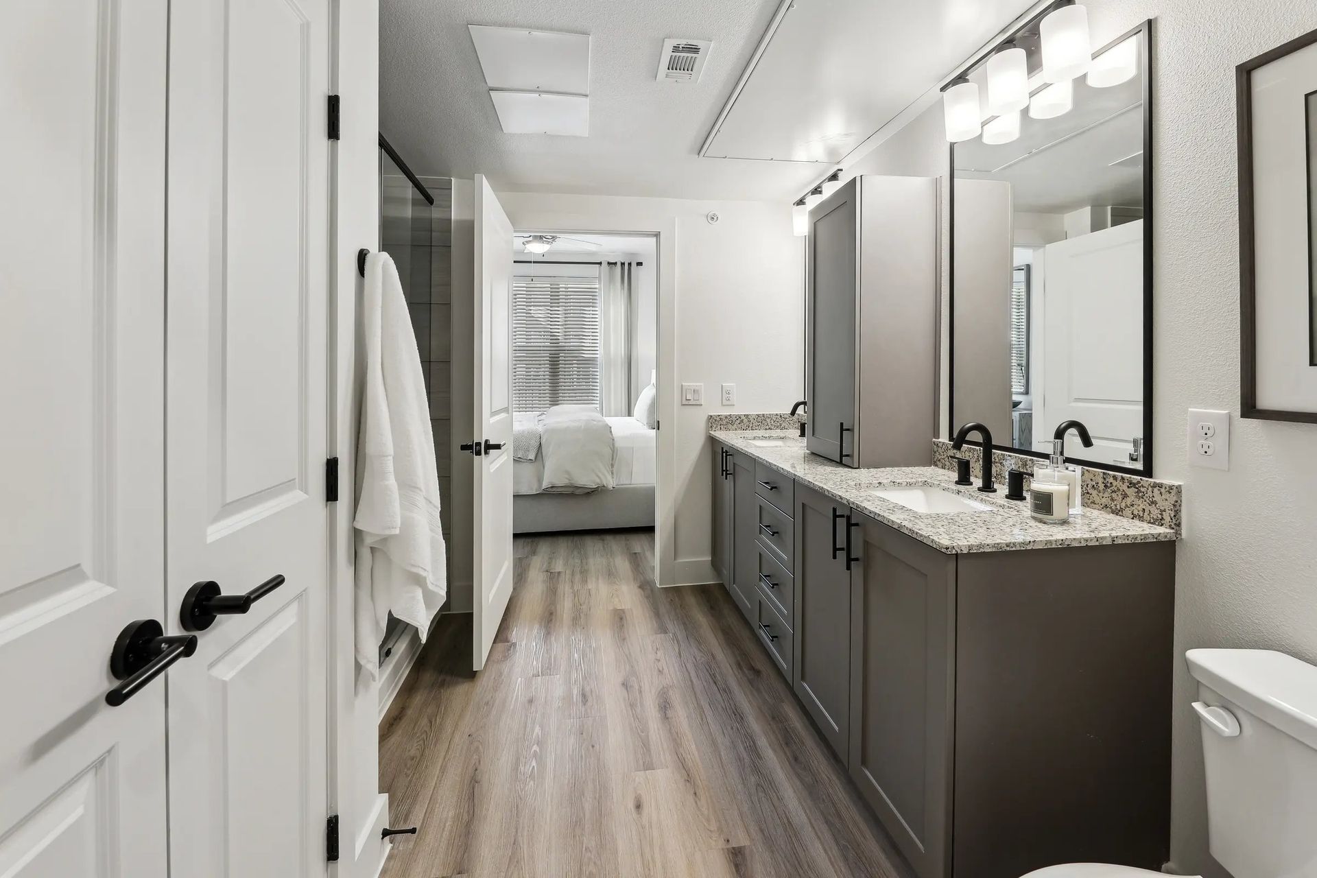 Bright modern bathroom with double vanity, mirrors, white walls, and wood-look floor leading to a bedroom.