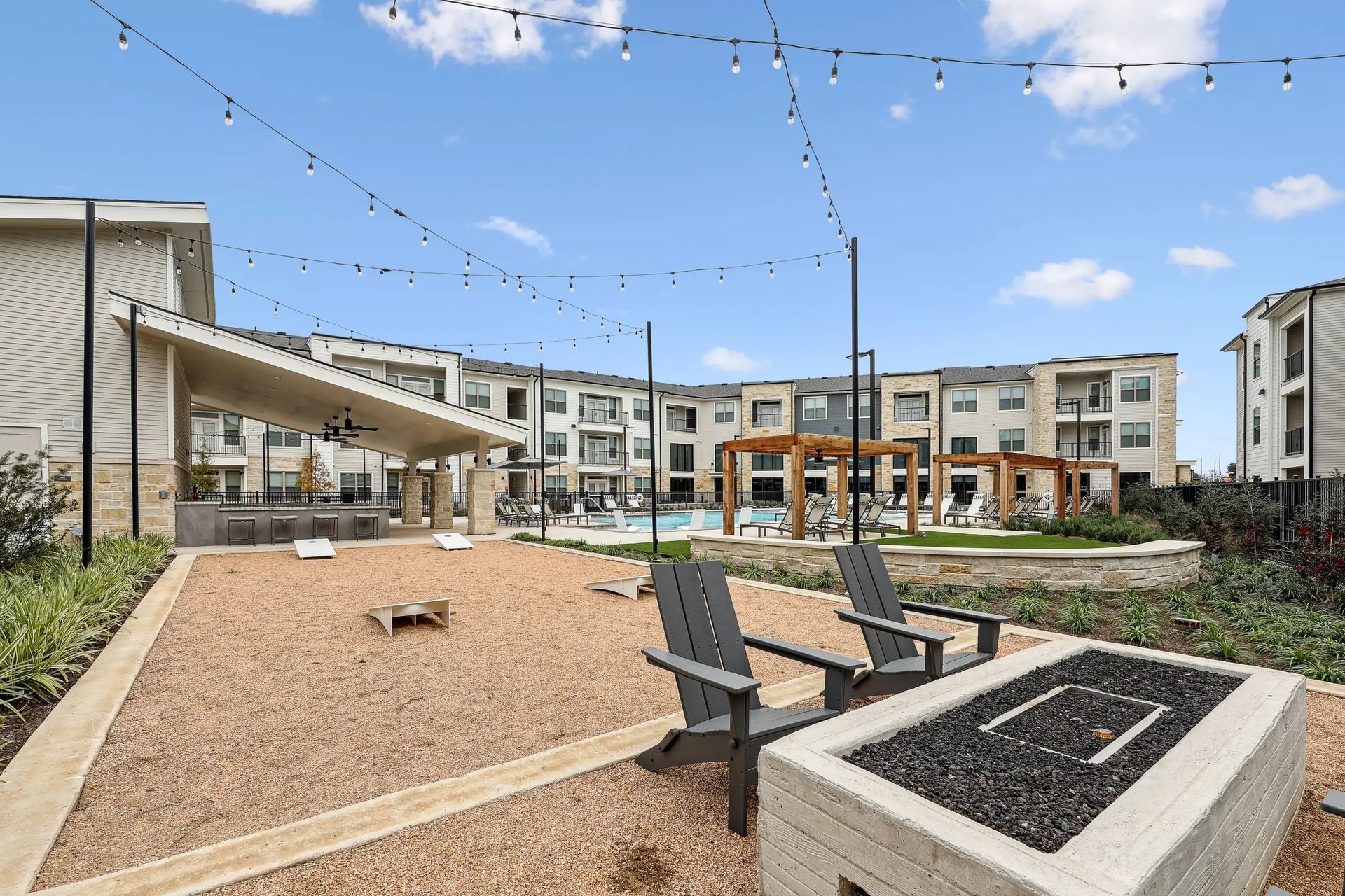 Modern apartment courtyard with benches, gravel seating area, fire pit, and string lights under blue sky