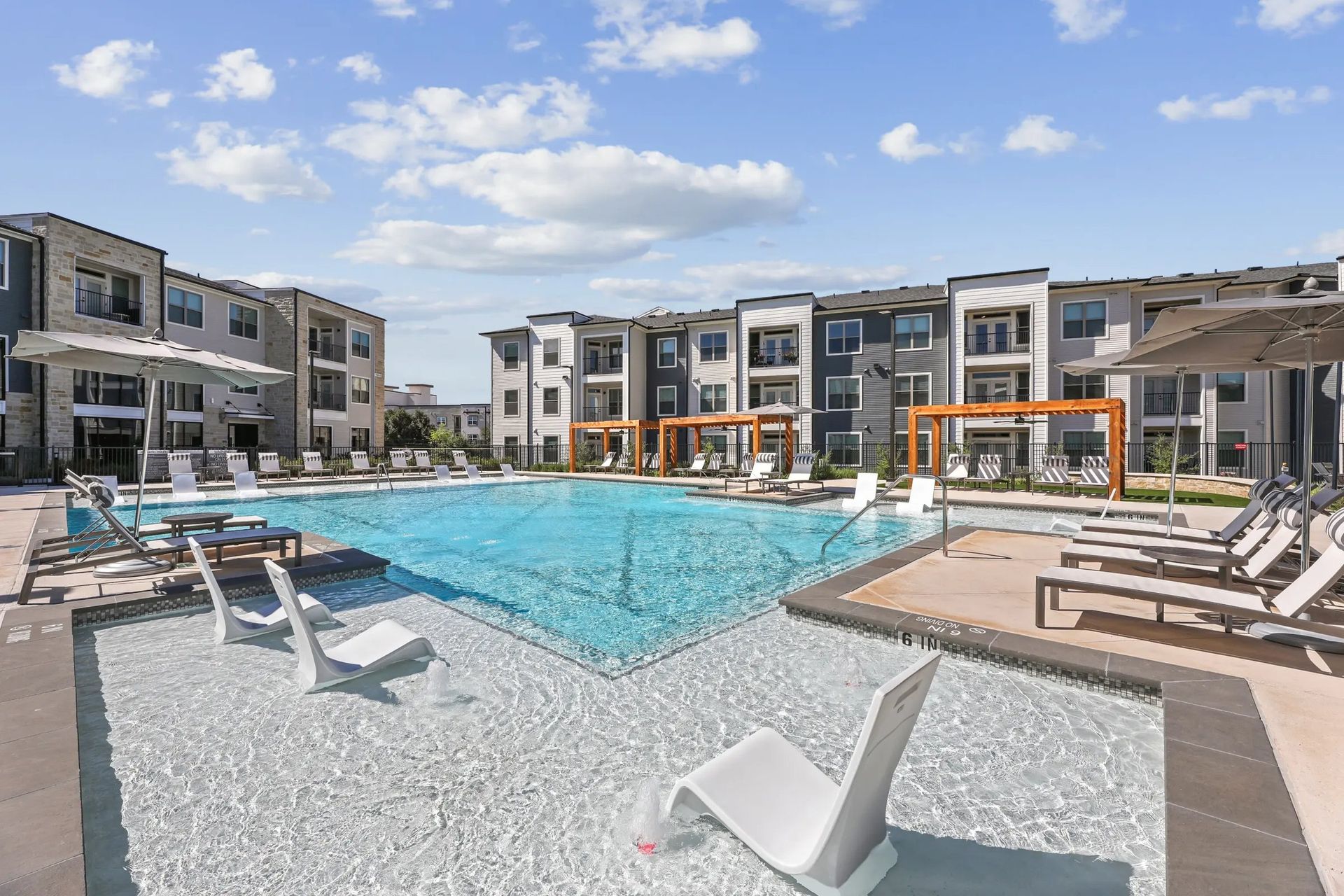 Modern apartment pool courtyard with loungers, lounge chairs, and blue water under a partly cloudy sky