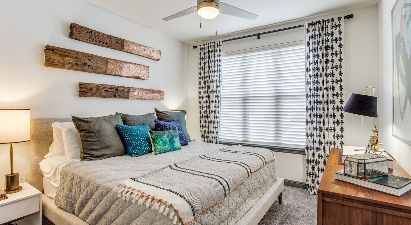 Bright bedroom with white bed, gray and blue pillows, wooden wall art, and window with patterned curtains