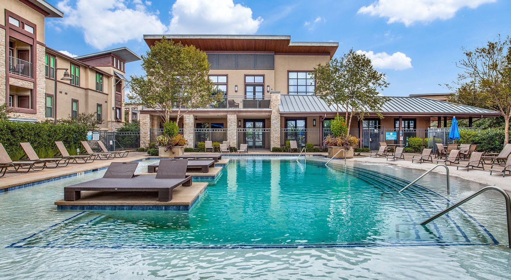 Modern apartment complex with a turquoise outdoor pool, lounge chairs, and clear blue sky