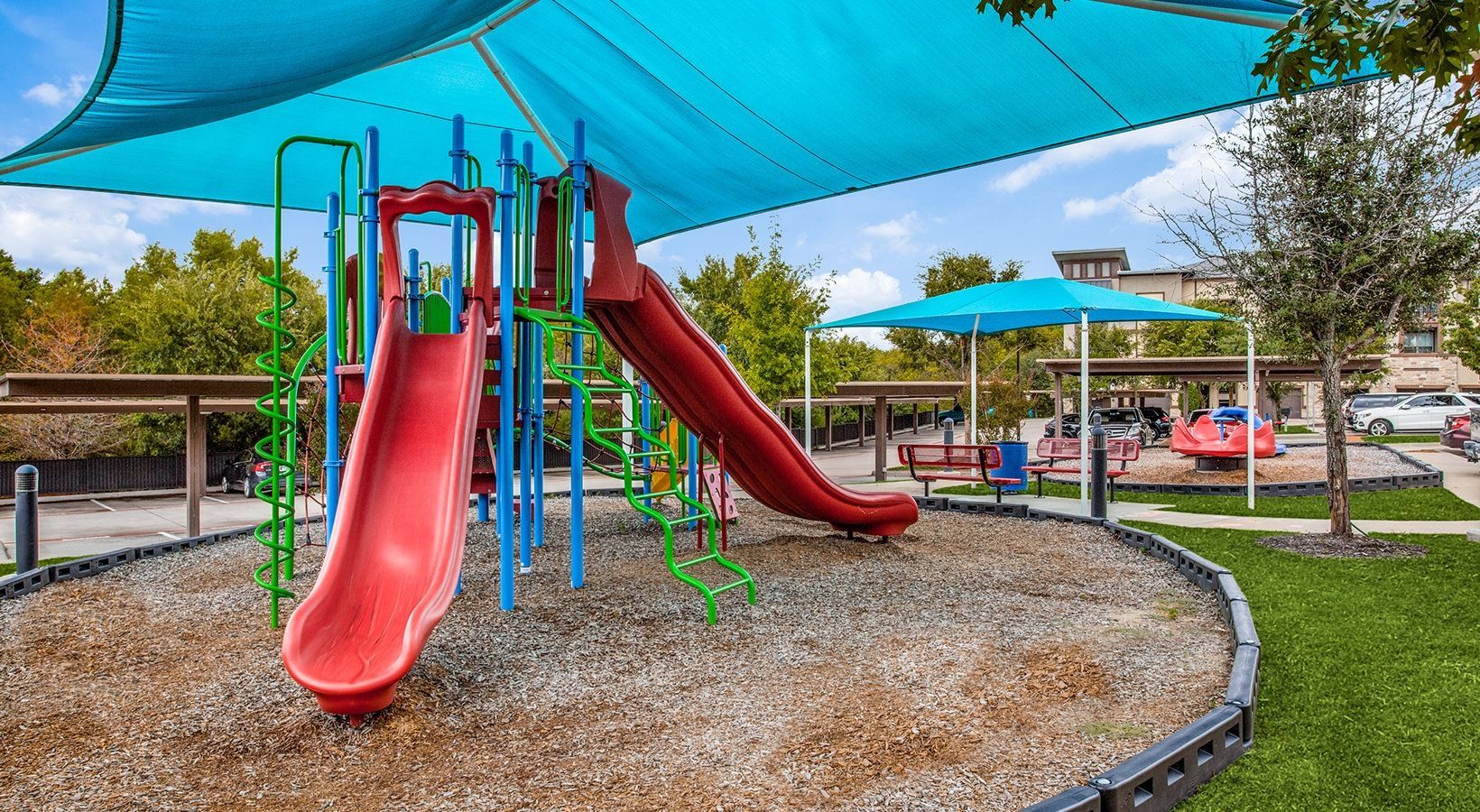 Playground with red slides and blue shade sails over a gravel play area