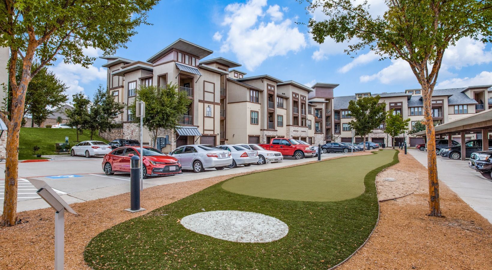 Apartment complex courtyard with parked cars, trees, and a landscaped lawn on a sunny day