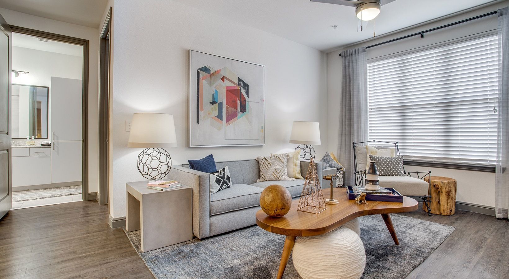 Bright modern living room with white furniture, abstract wall art, gray rug, and a wooden coffee table by a window.