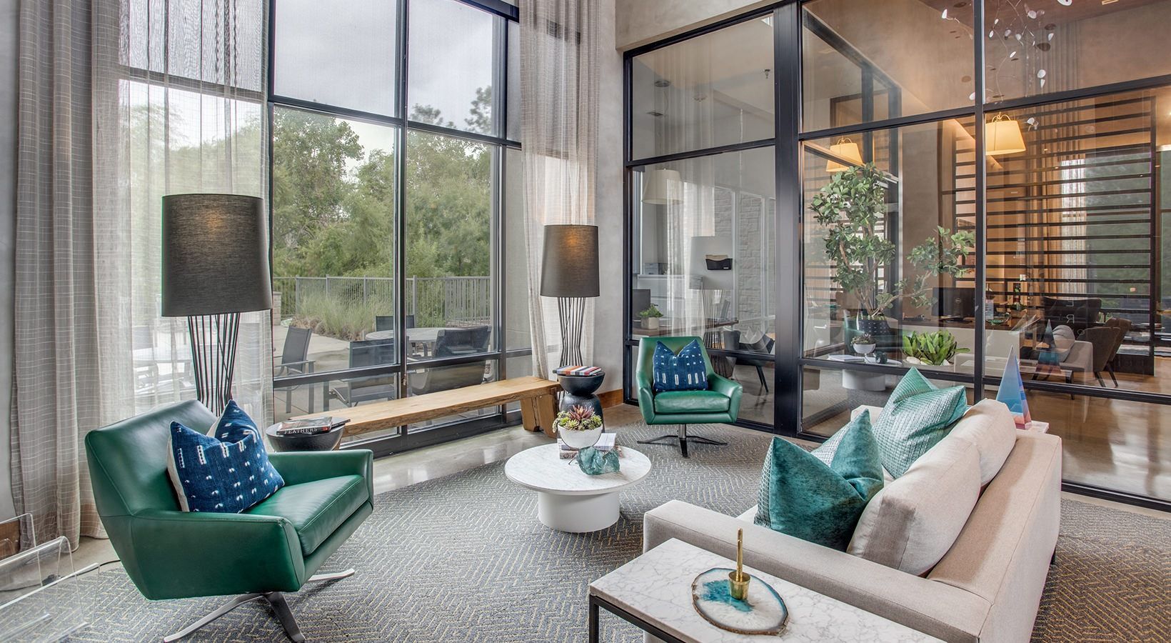 Modern living room with tall windows, teal chairs, beige sofa, and a staircase.