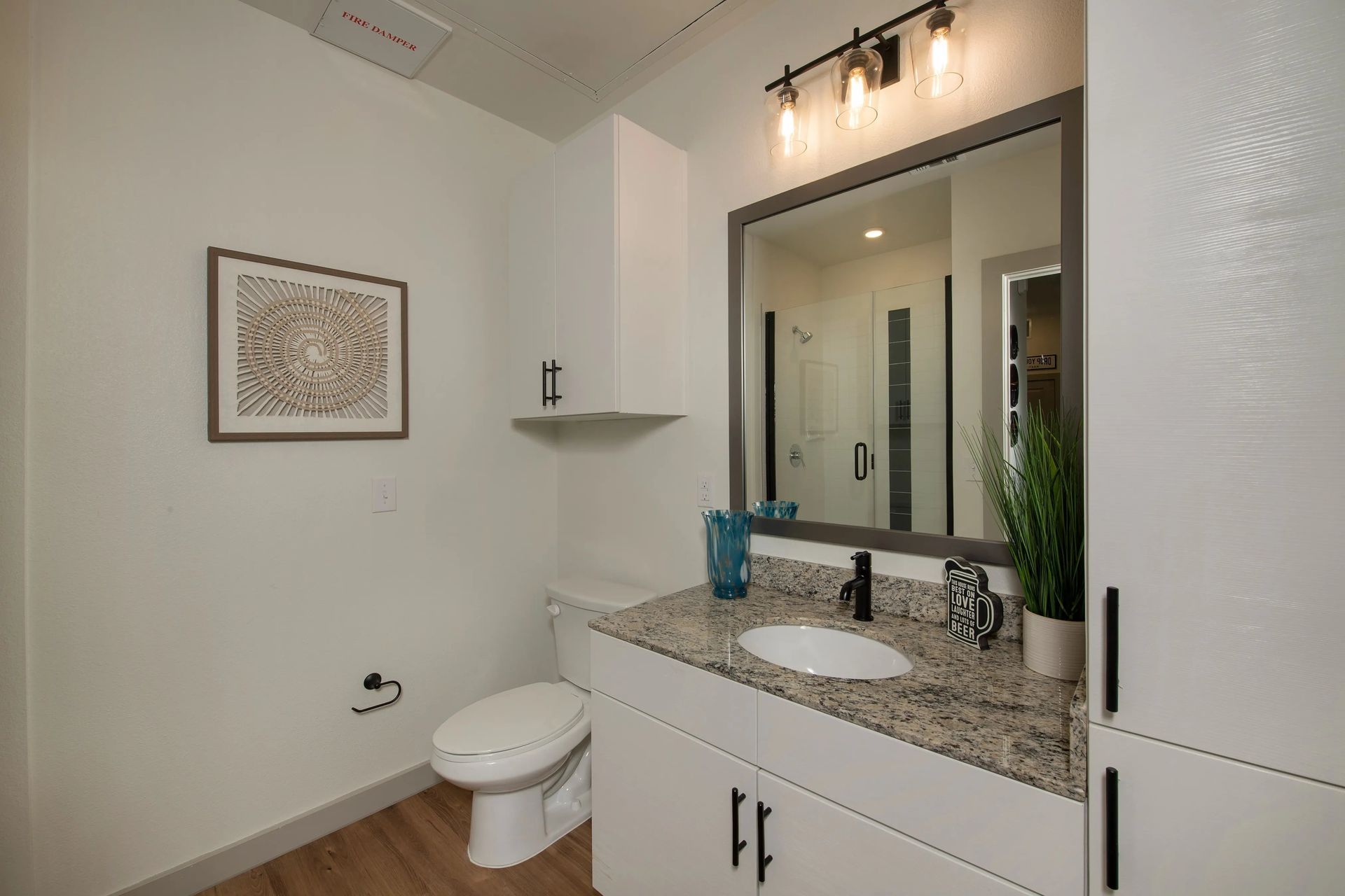 Modern bathroom vanity with granite countertop, mirror, wall art, and toilet in a bright neutral room