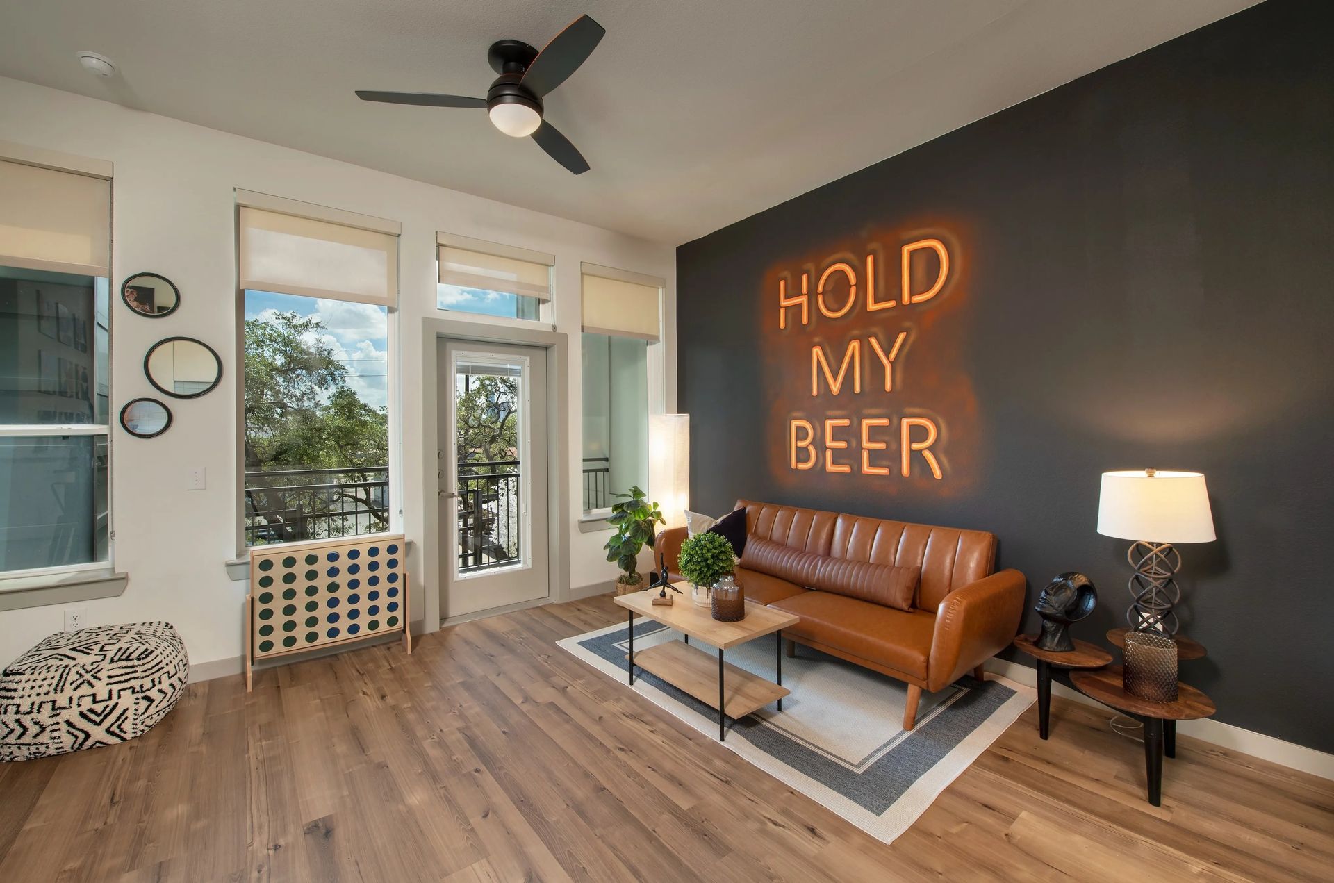 Cozy living room with neon “HOLD MY BEER” sign, leather sofa, coffee table, and large windows.