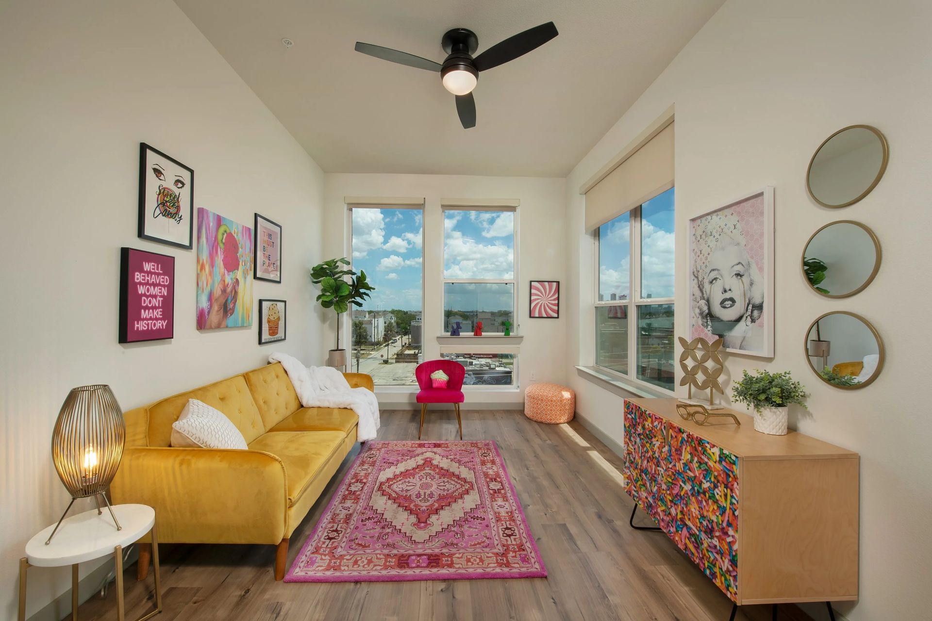 Bright living room with yellow sofa, pink rug, colorful wall art, and a large window with city view