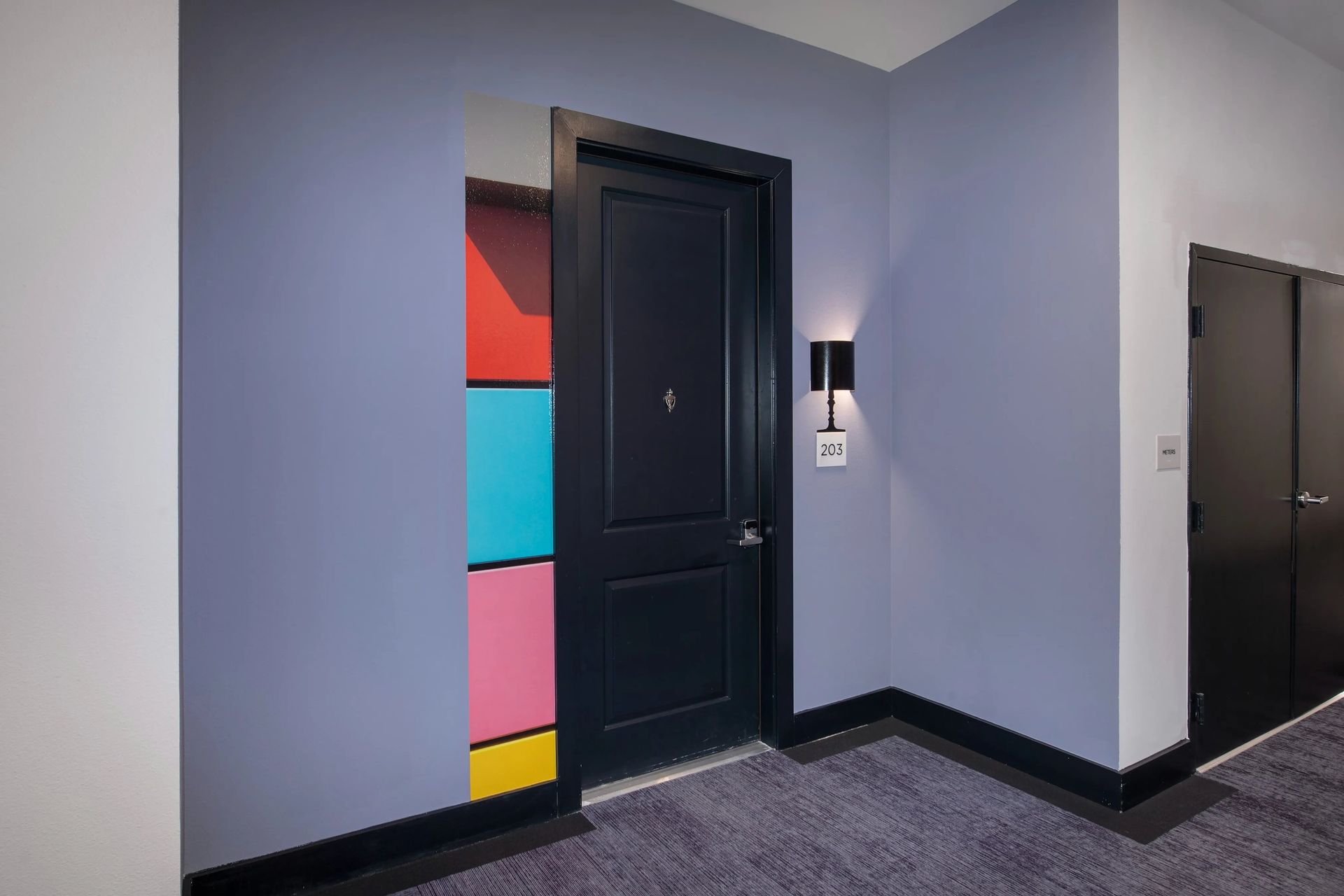 Hallway with a black door beside colorful wall panels and a wall sconce.
