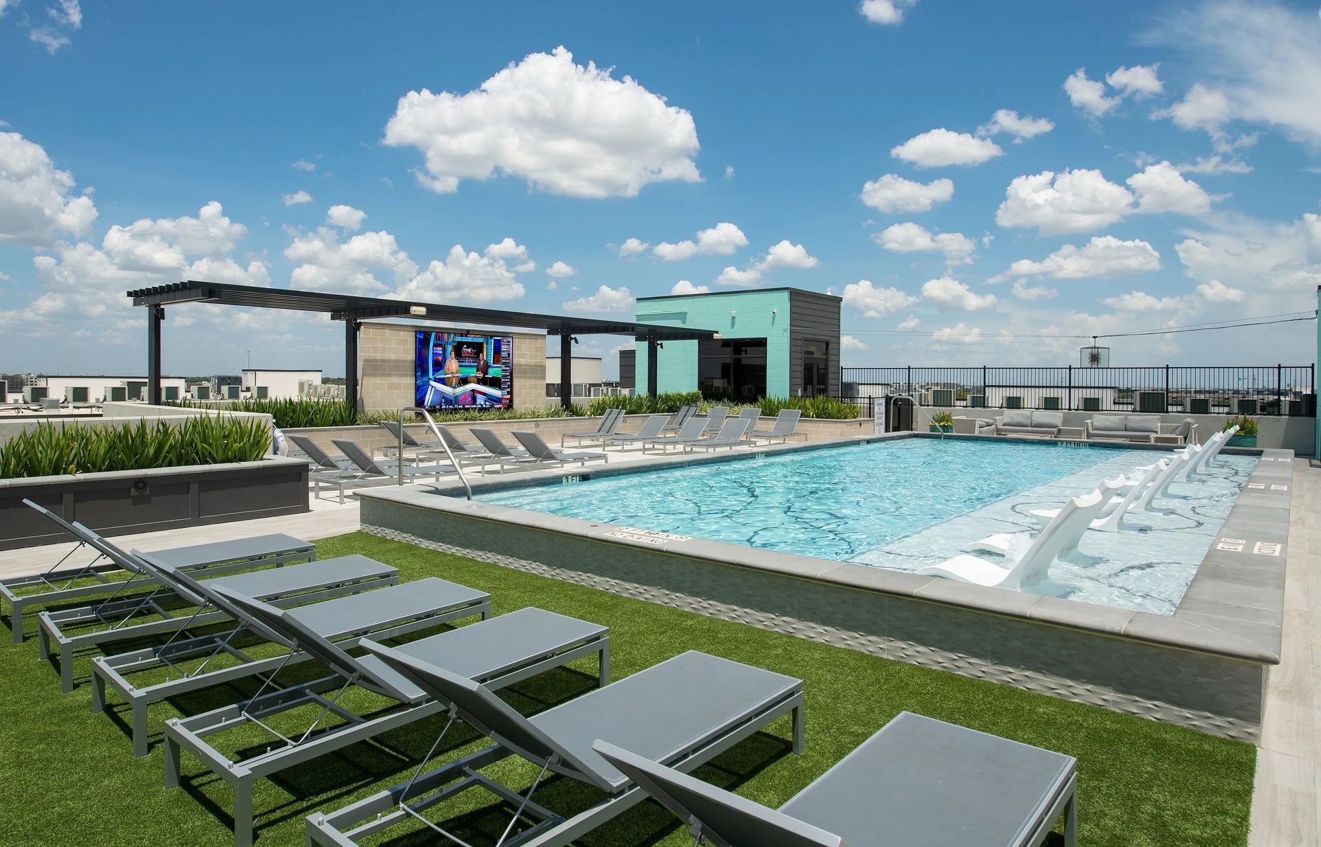 Rooftop pool with lounge chairs, grassy deck, and modern building under a bright blue sky