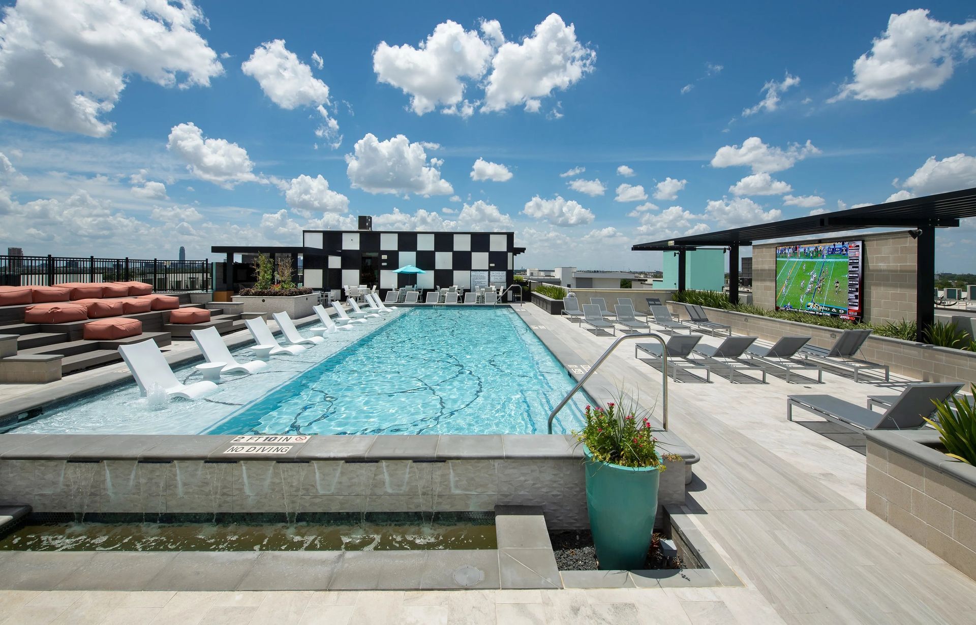 Outdoor rooftop pool with lounge chairs, umbrellas, and a blue sky with clouds