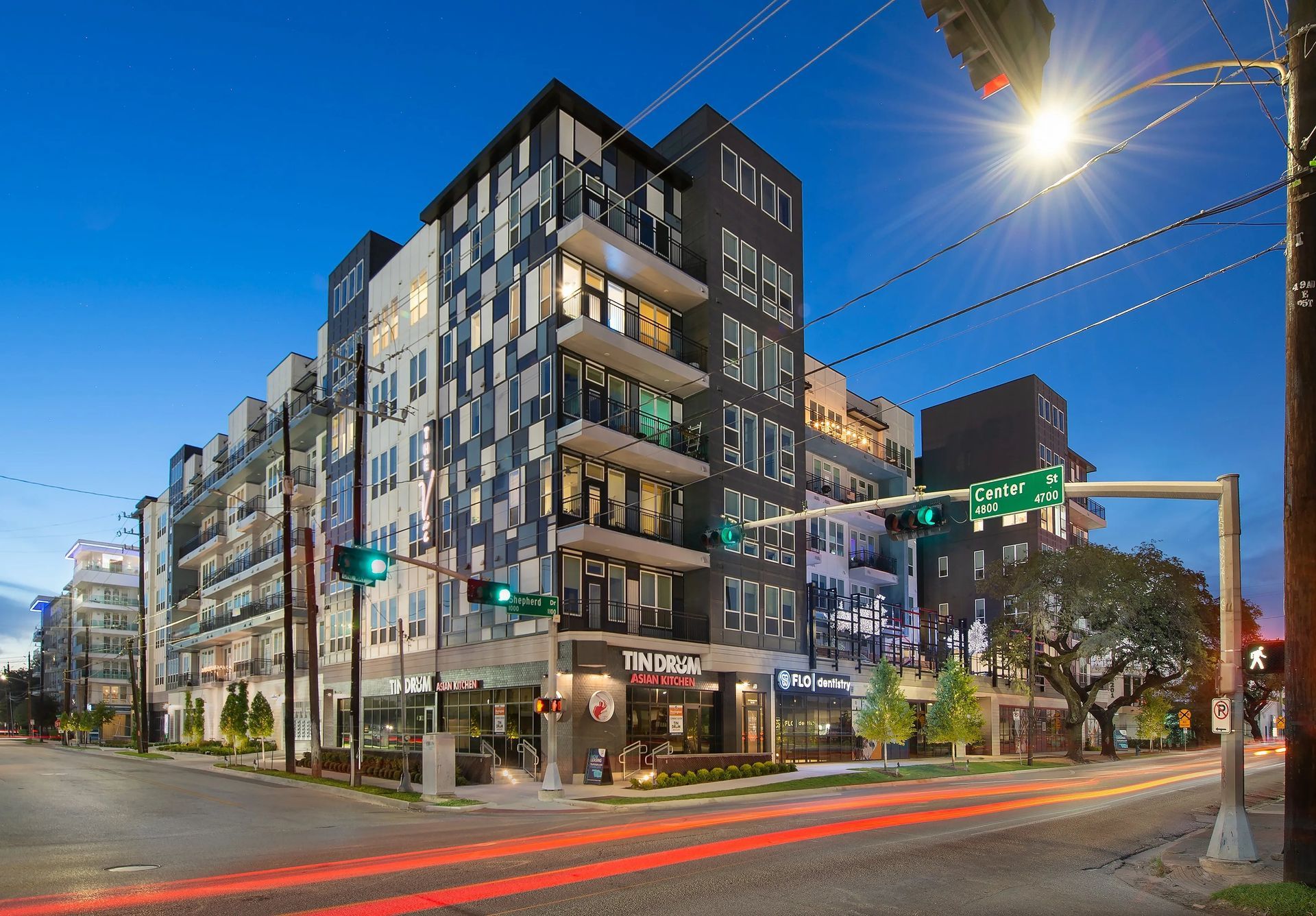 Modern apartment building at a city intersection at dusk, with light trails from passing cars.