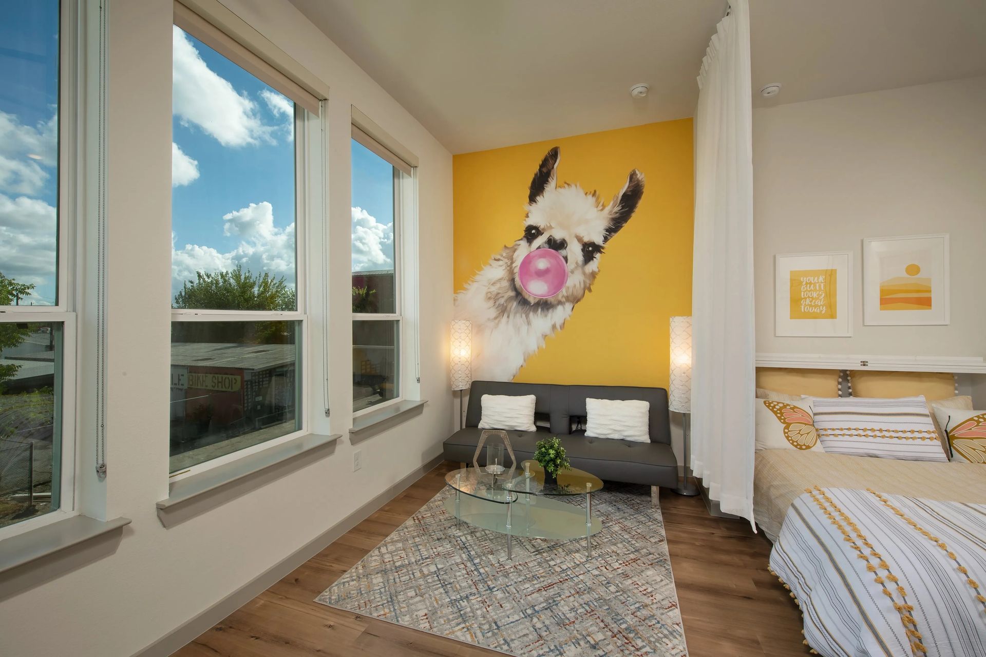 Bright bedroom with large windows, yellow accent wall art, bed, desk, and patterned rug