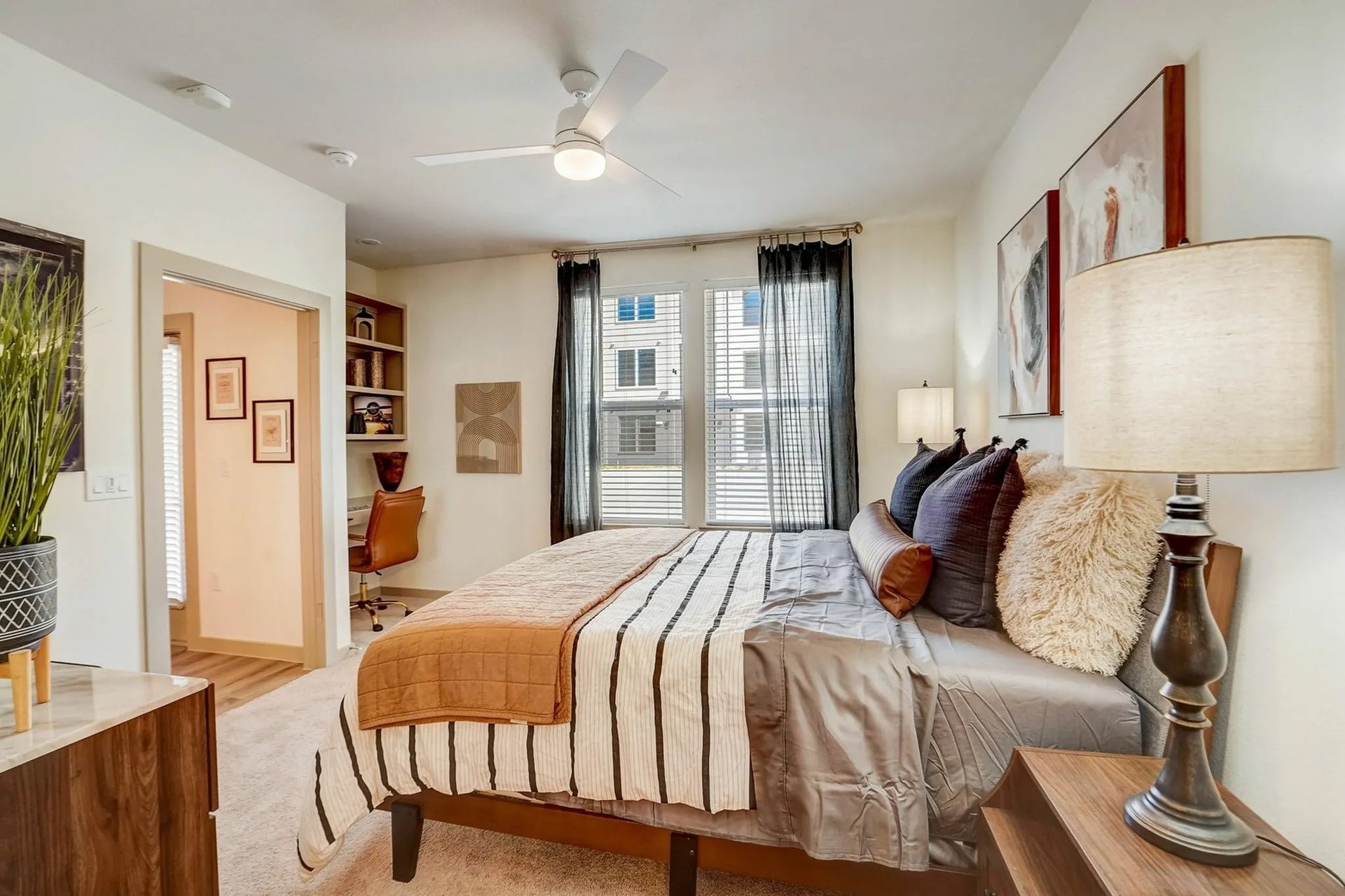 Bright, modern bedroom with a made bed, bedside lamp, ceiling fan, and window view of apartment buildings