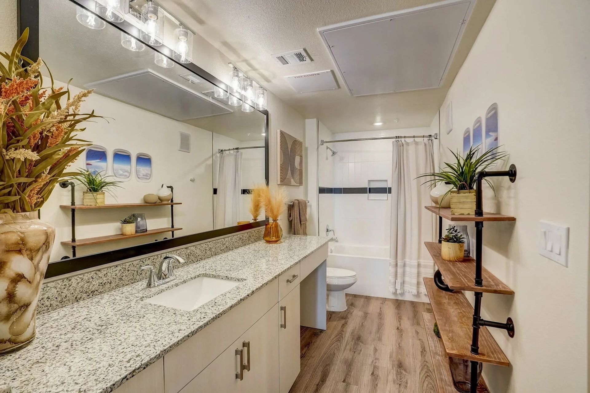 Bright bathroom with double vanity, granite countertop, mirror lights, and wood floor leading to a toilet area