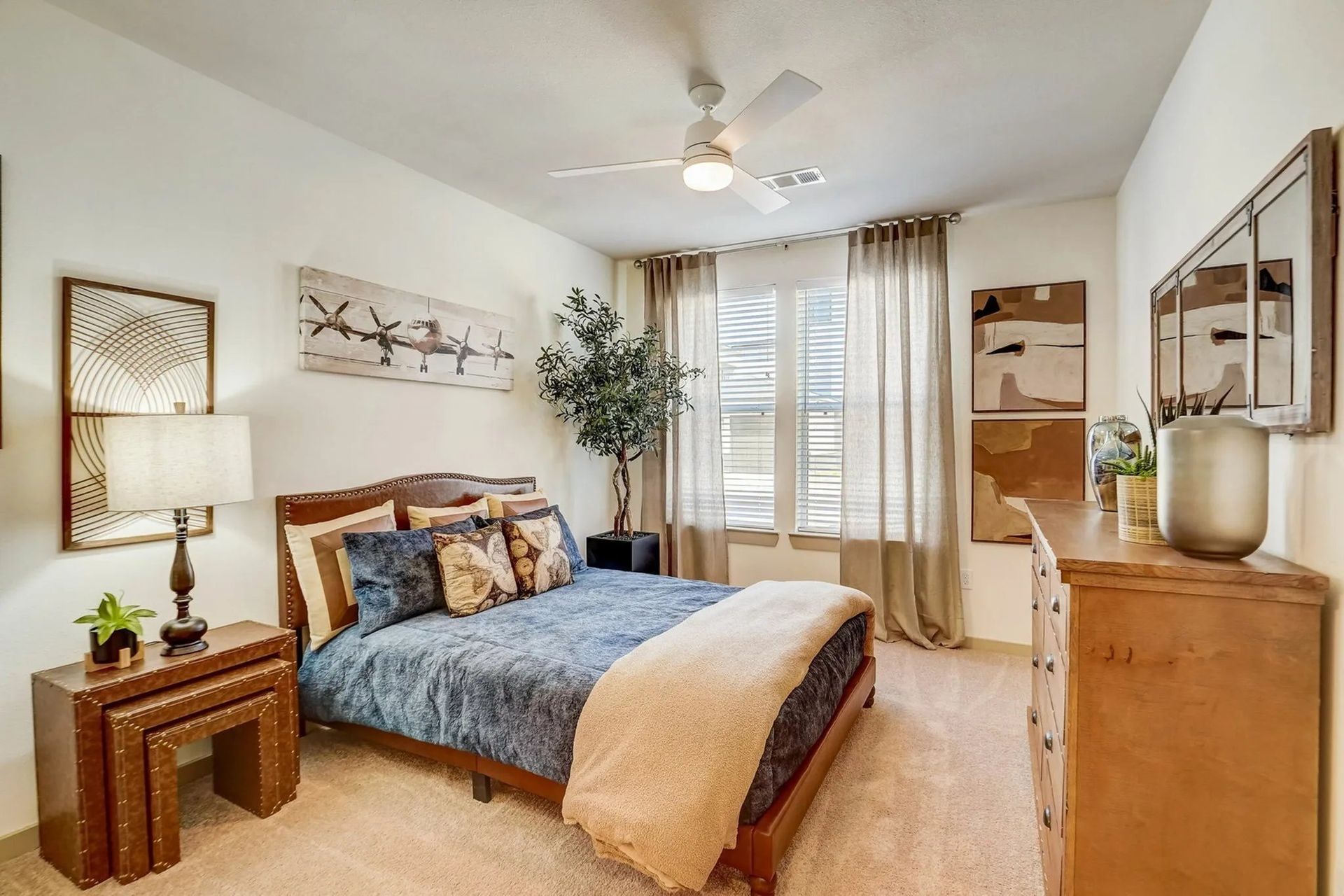 Bright bedroom with bed, nightstands, dresser, ceiling fan, and large window with curtains