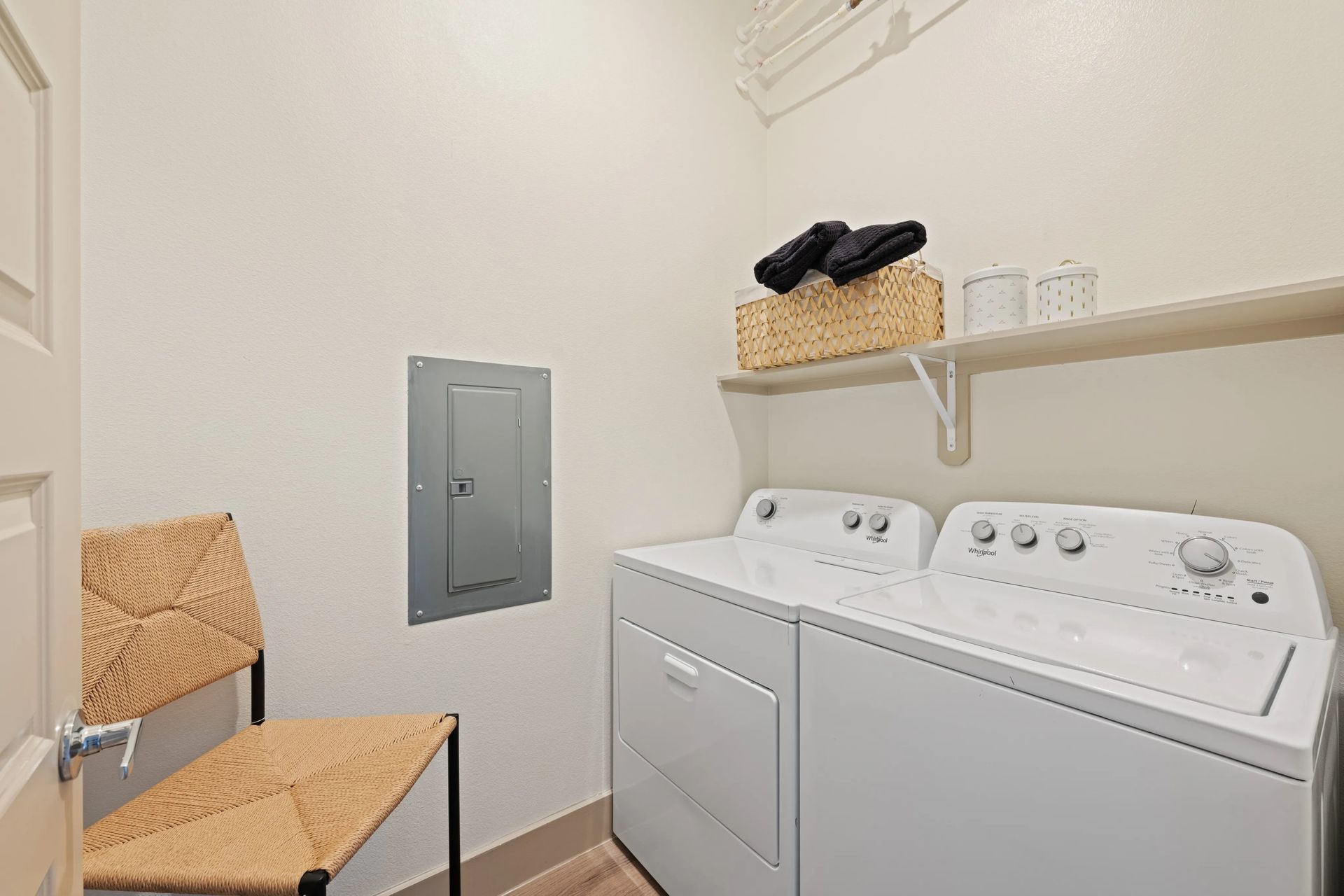 Laundry room with washer and dryer, woven chair, shelf with basket, and gray electrical panel