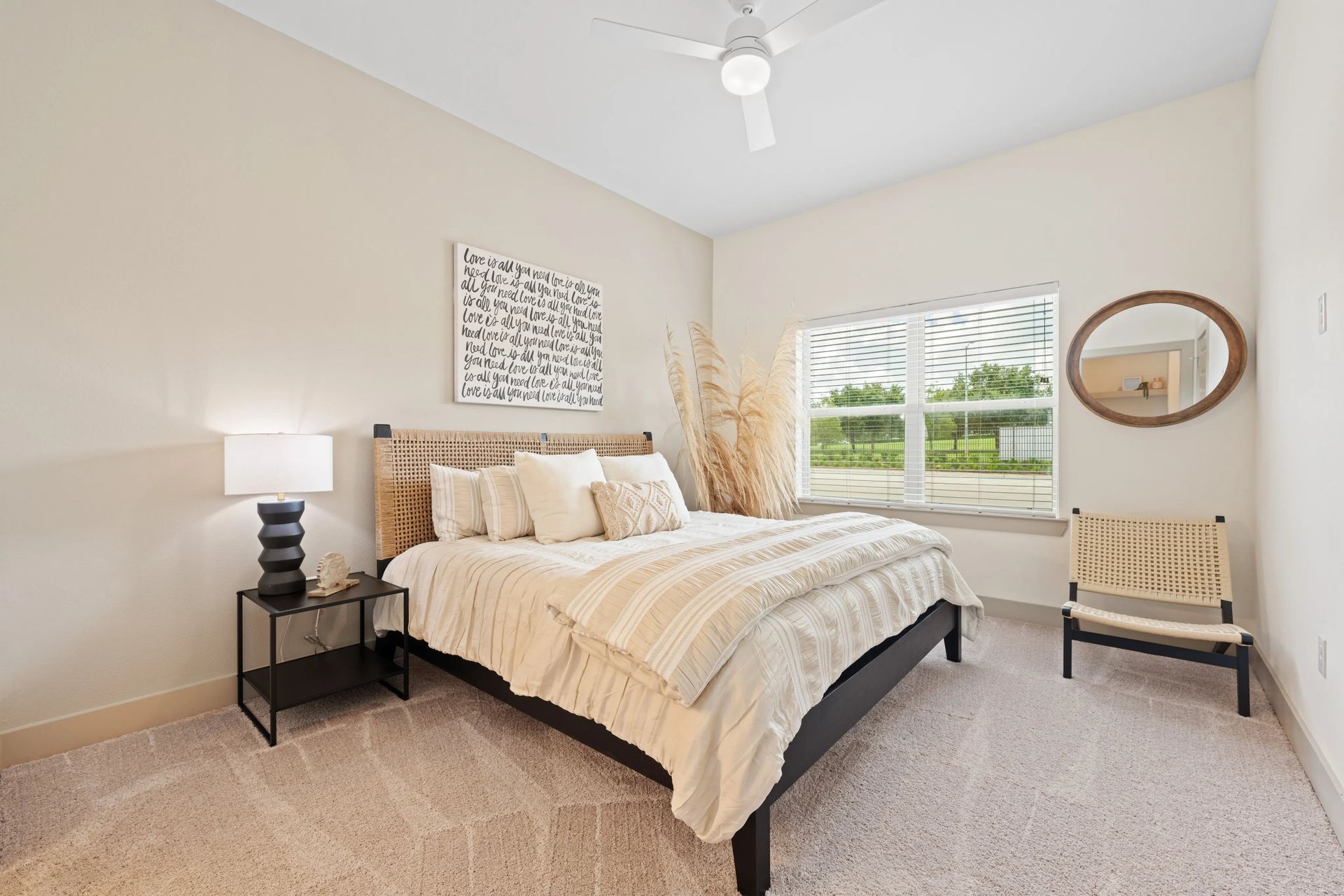Bright bedroom with a neatly made bed, beige walls, window view, and a small chair and lamp.