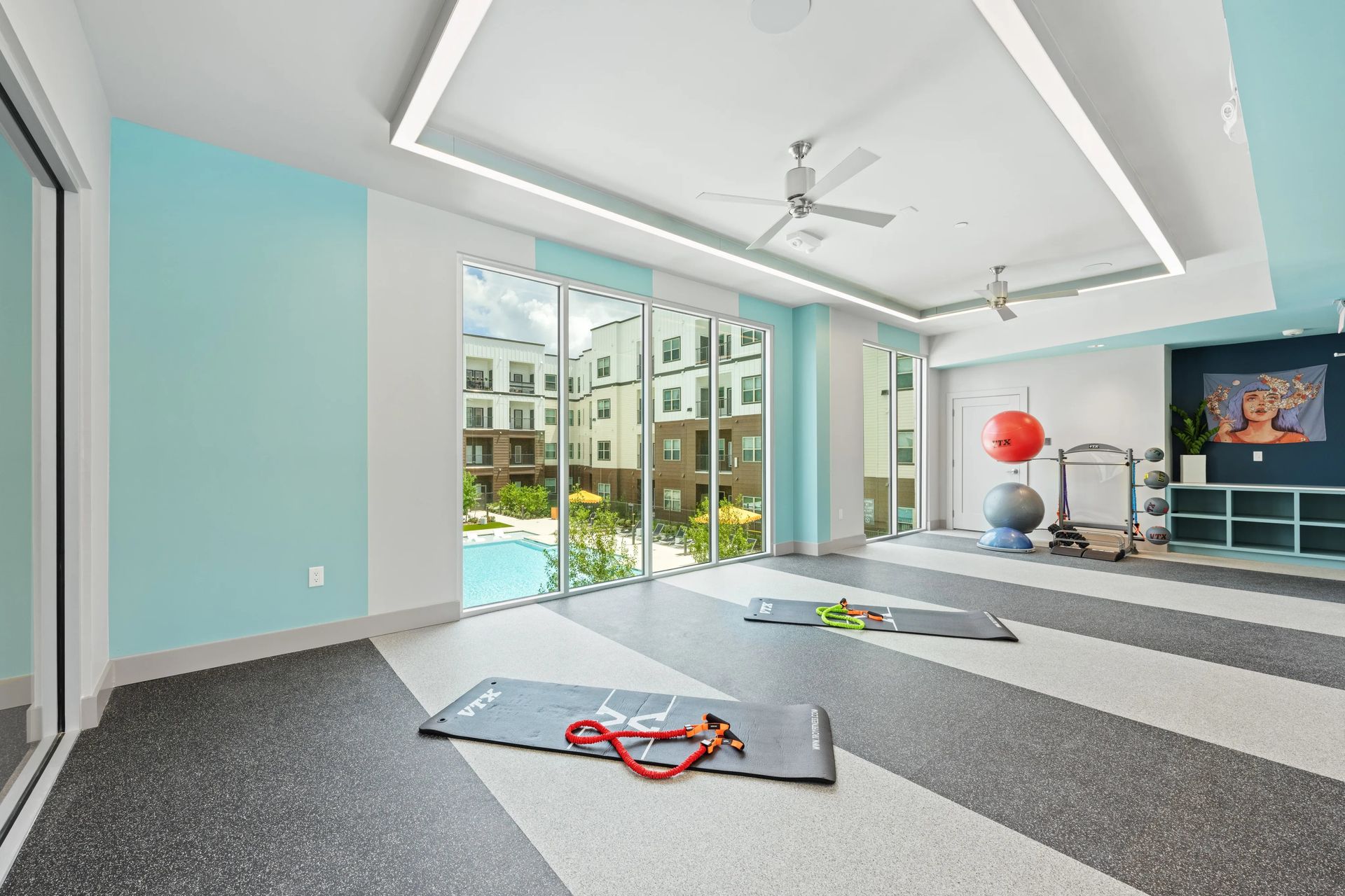 Spacious fitness room with mats, exercise equipment, and large windows overlooking a pool and apartment courtyard