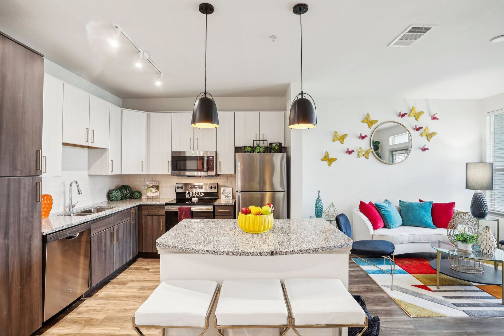 Bright open kitchen and living room with white cabinets, gray sofa, colorful pillows, and yellow wall butterflies.