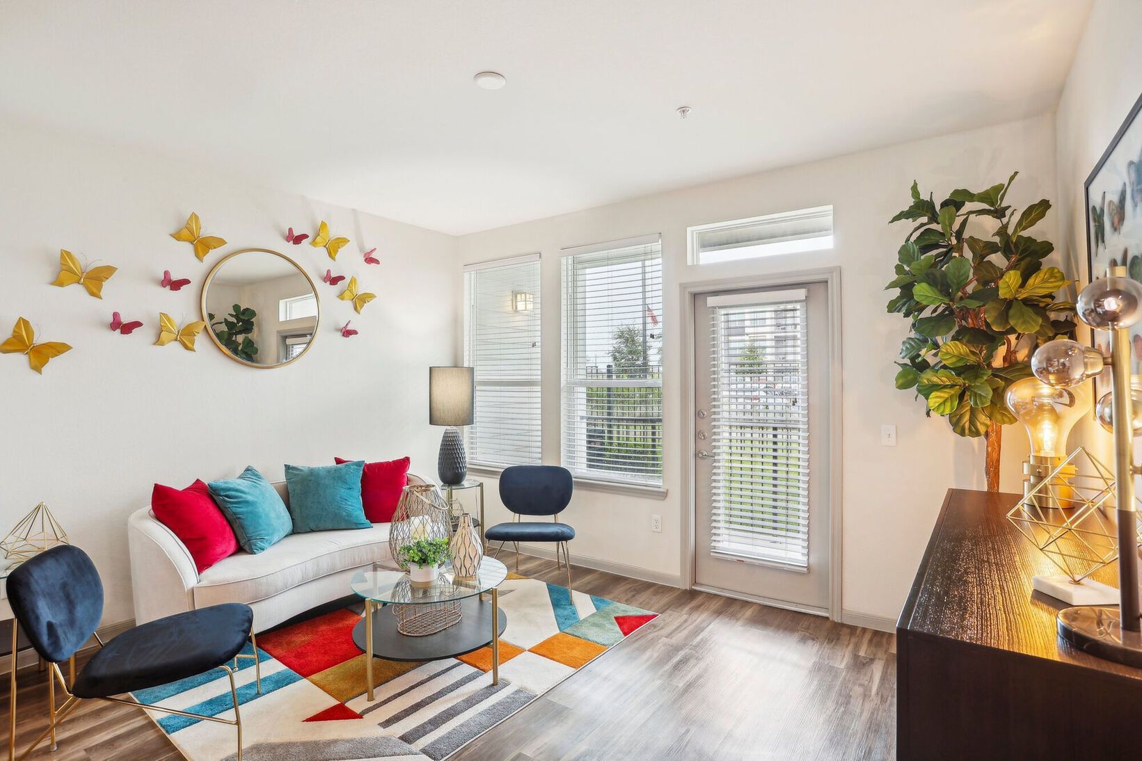 Bright living room with white sofa, colorful pillows, patterned rug, round mirror, and flowers by the window