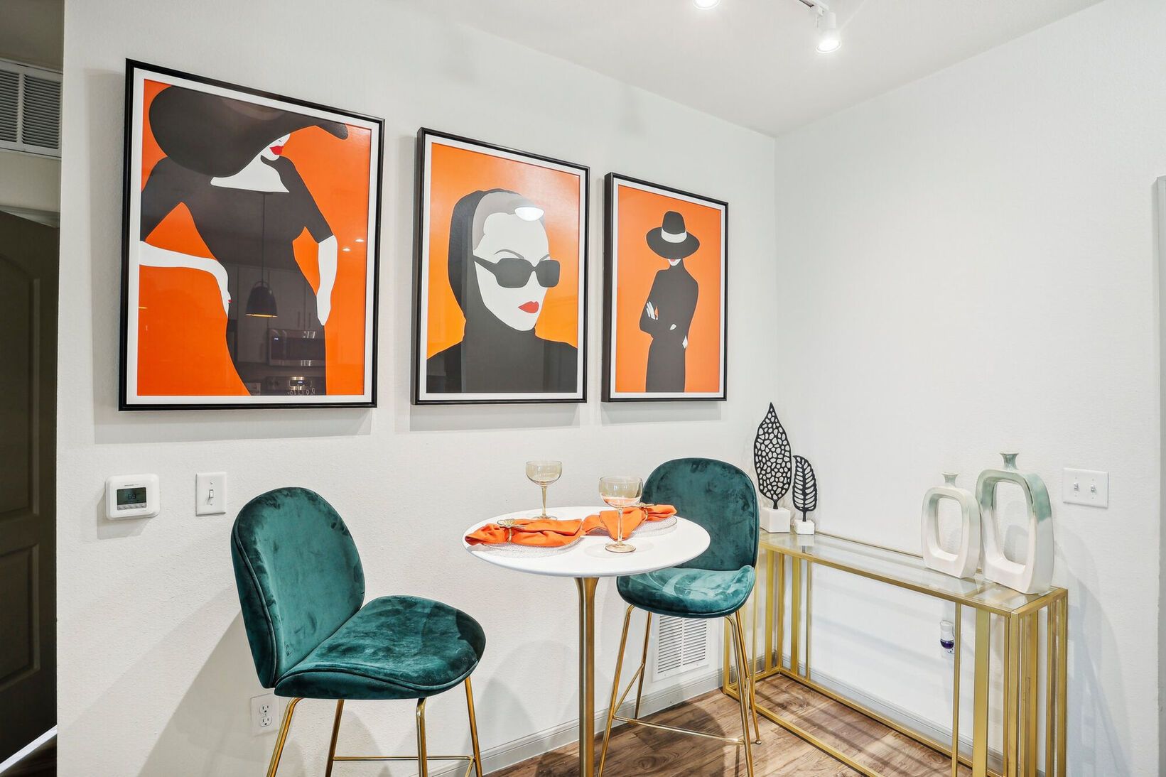Modern dining nook with teal chairs, round table, orange abstract art, and gold console against a white wall
