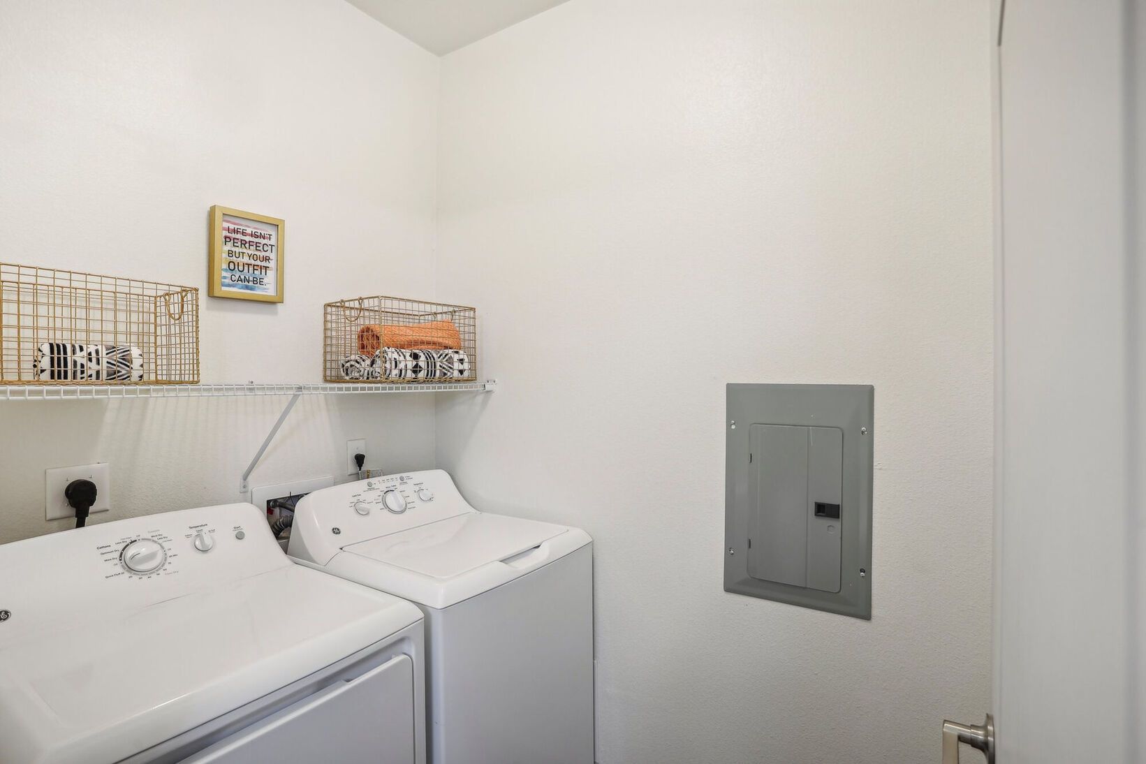 Laundry room with two white washers, wall shelves, and a gray electrical panel on a white wall