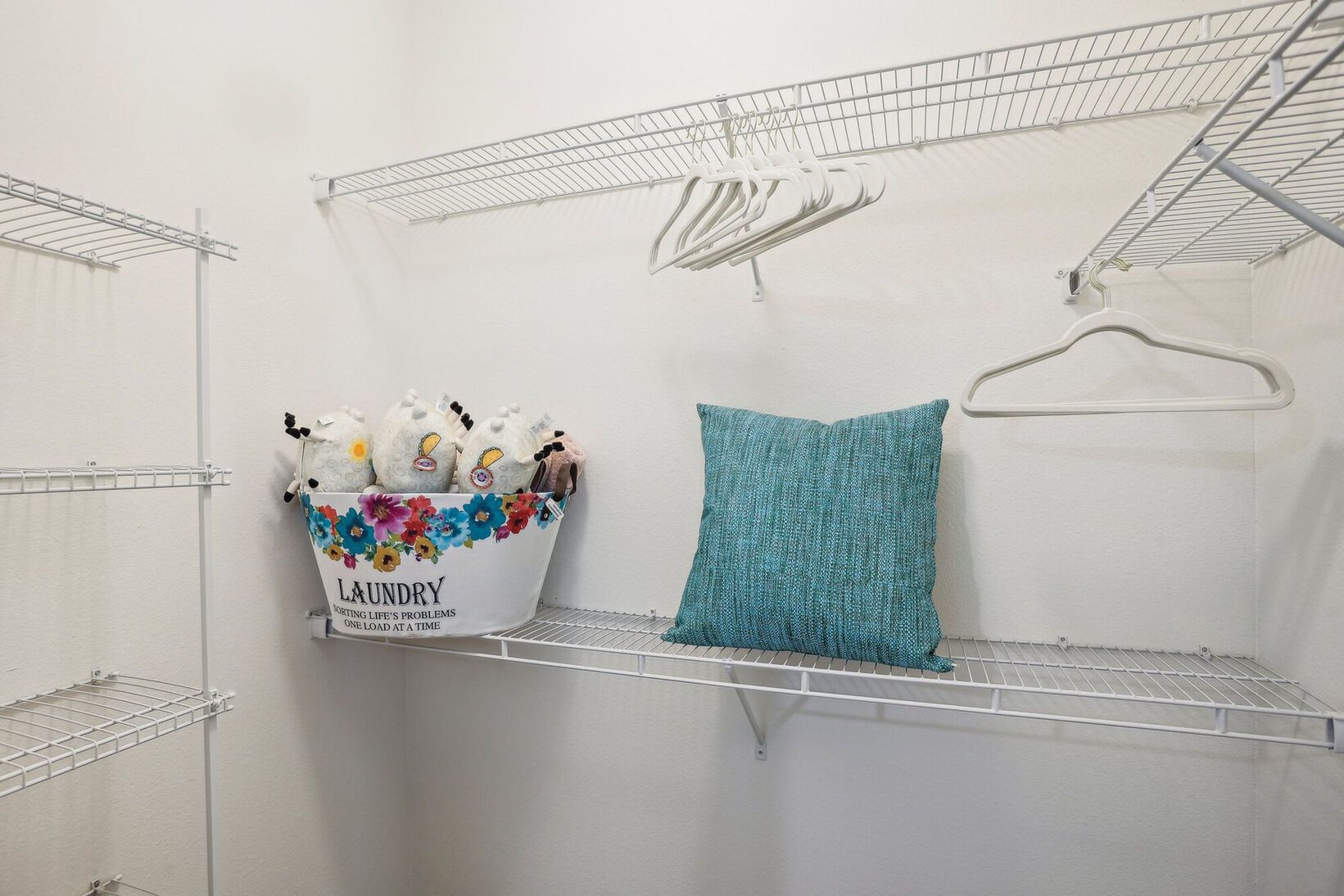 Closet shelf with a basket of stuffed animals, teal pillow, and wire hangers on white shelves.