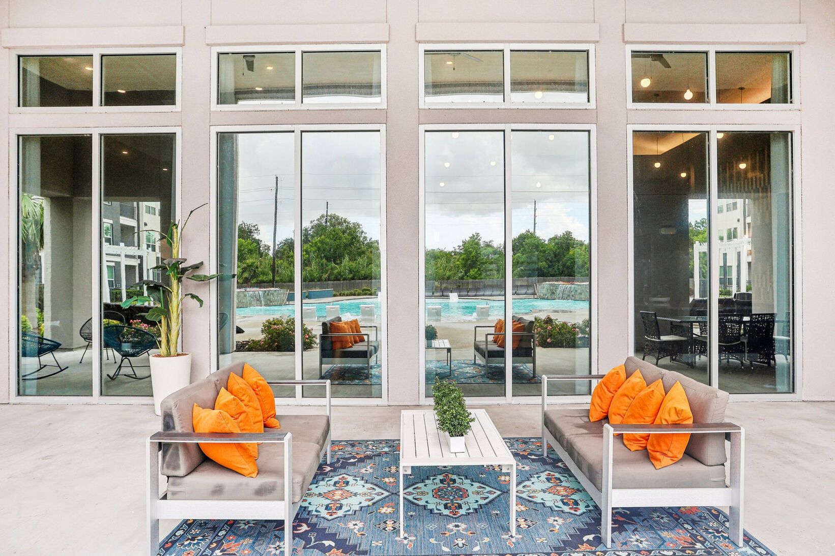 Modern patio with glass doors, two white chairs with orange pillows, and a pool view outside