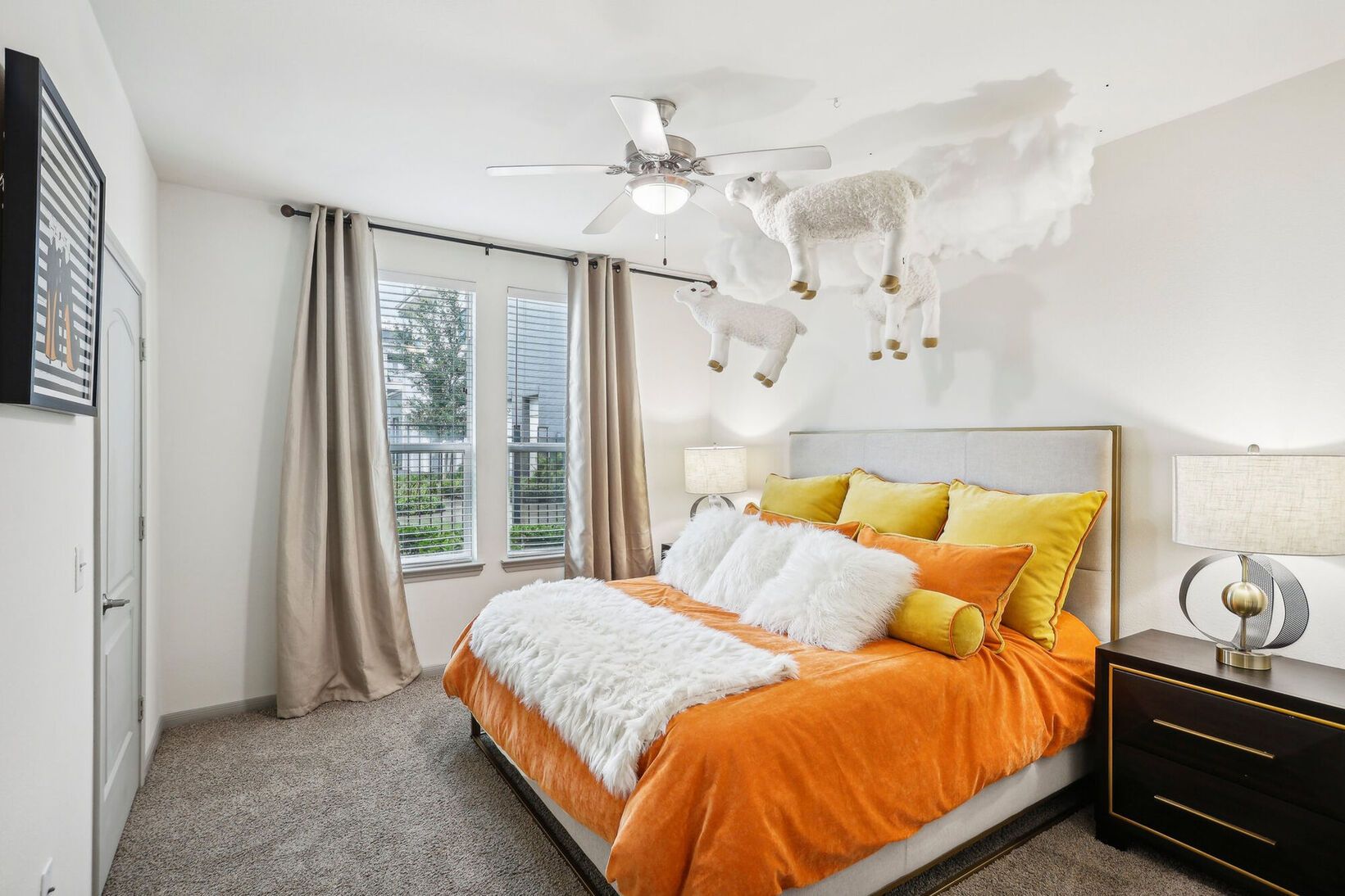Bright bedroom with orange bedspread, white pillows, gray carpet, and large windows with curtains.