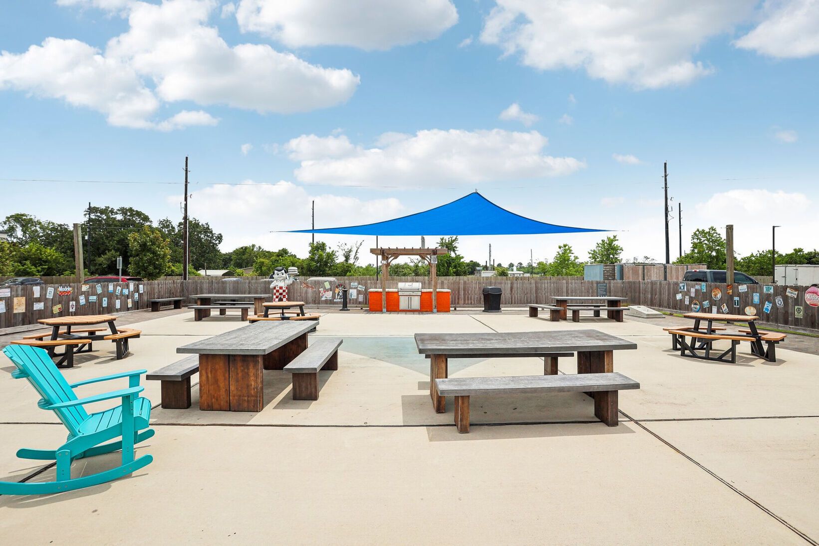 Outdoor patio with picnic tables and benches under a blue shade sail on a sunny day