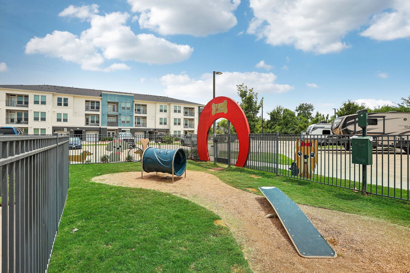 Playground with a red arch, blue tunnel, and ramp on grass beside apartment buildings