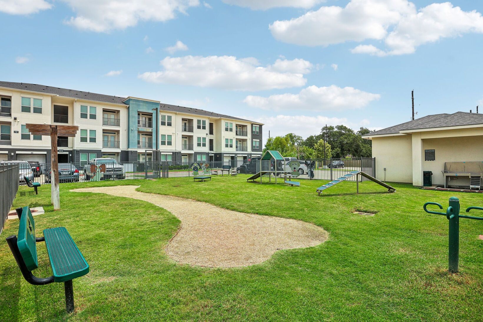 Apartment courtyard with grassy lawn, gravel path, benches, and playground equipment under a blue sky