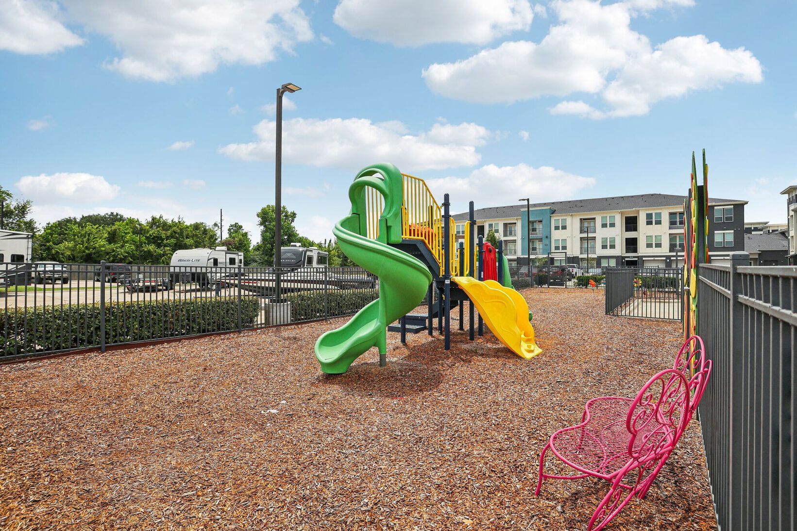 Colorful playground with green slide, yellow ramp, and pink climbing net on wood chips under a blue sky