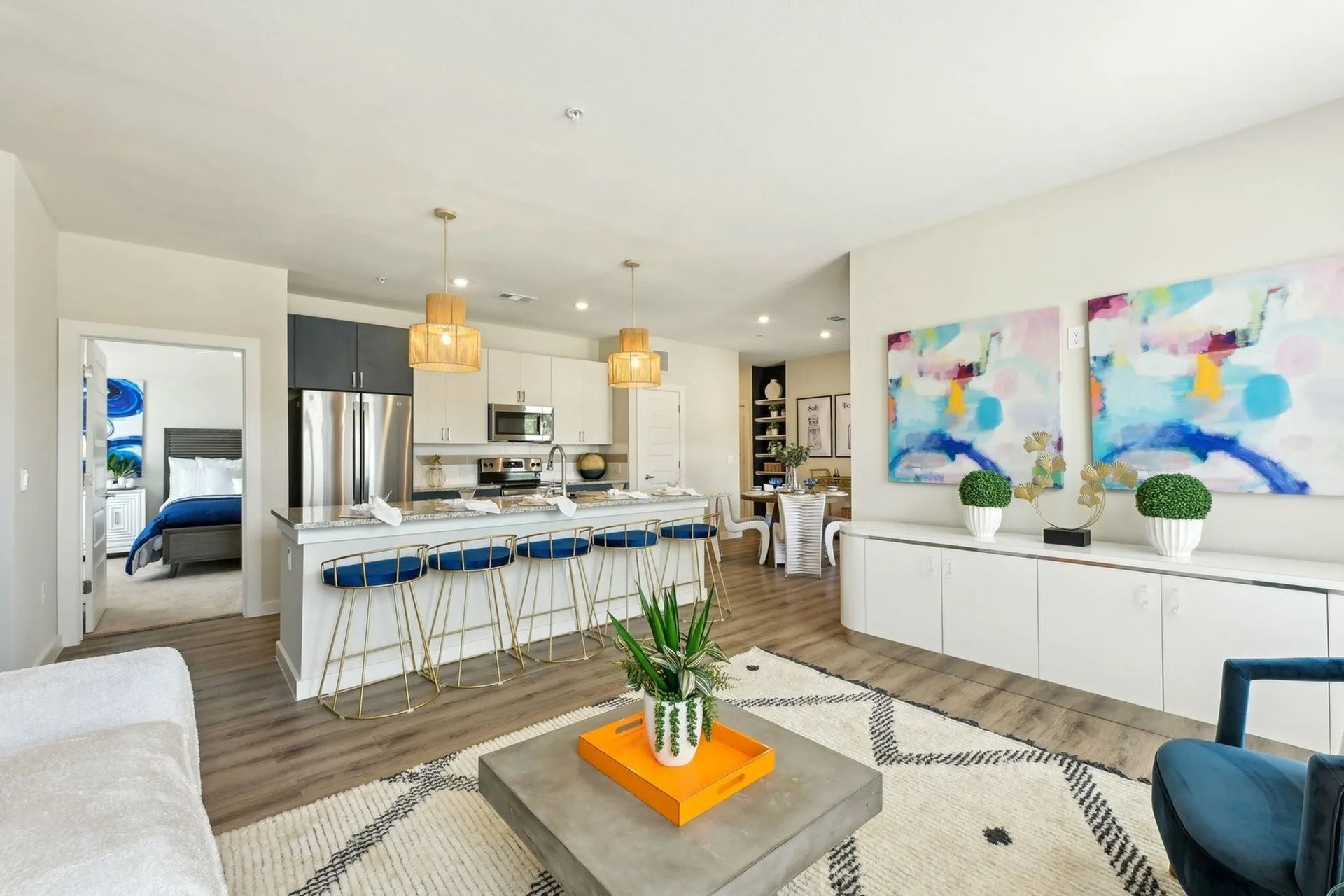 Bright modern living room with open kitchen, blue bar stools, colorful wall art, and coffee table centerpiece