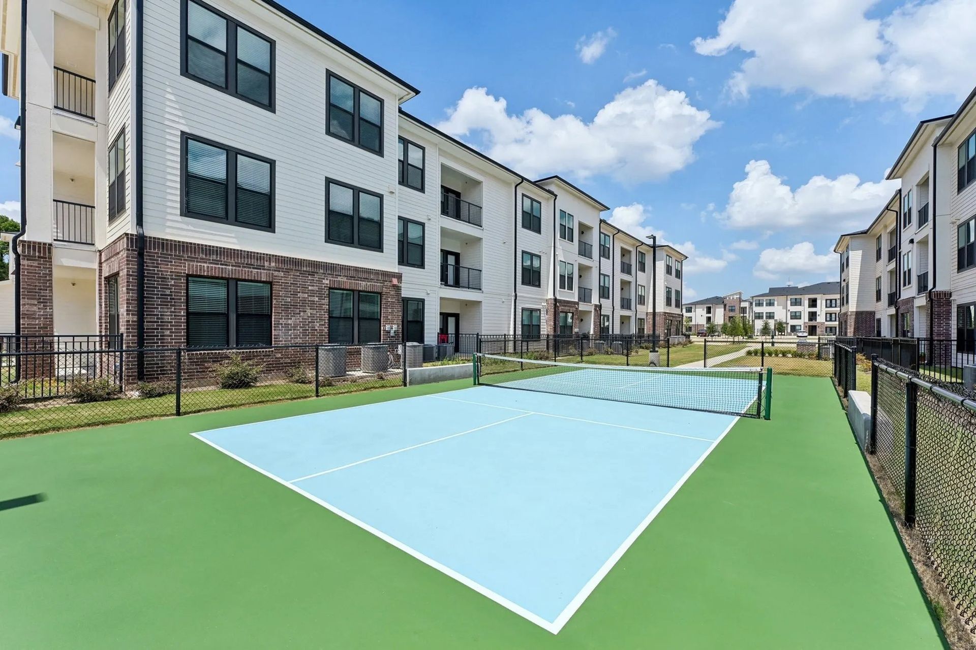 Apartment courtyard with a blue outdoor court, green turf, and modern buildings under a partly cloudy sky