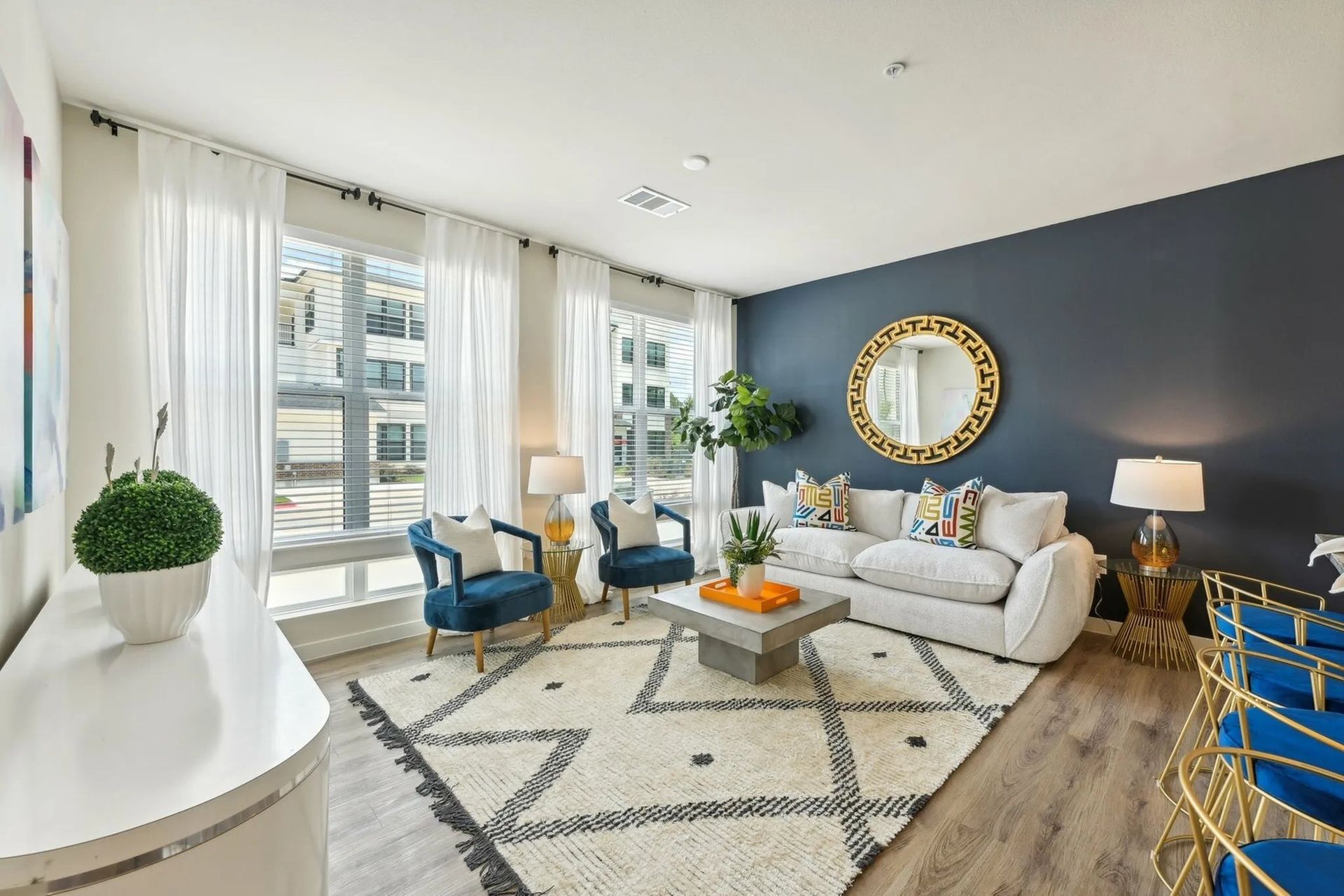 Bright modern living room with white sofa, blue chairs, large windows, and gold mirror on dark accent wall