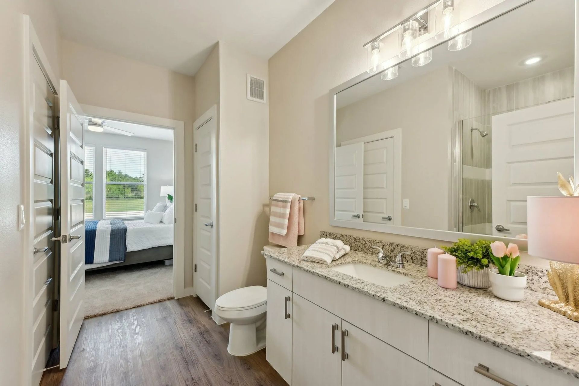 Bright bathroom vanity with granite countertop, large mirror, and open doorway to a bedroom with a bed and window.