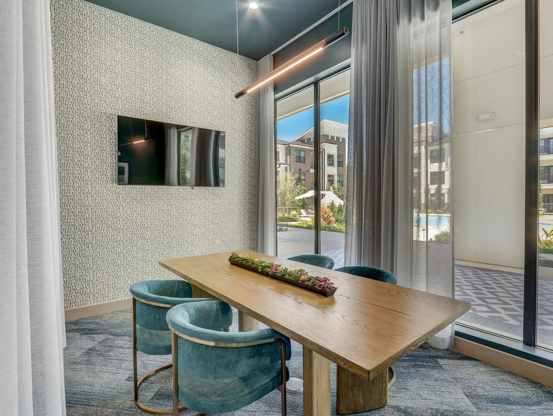 Modern meeting room with wooden table, teal chairs, wall-mounted TV, and floor-to-ceiling glass doors