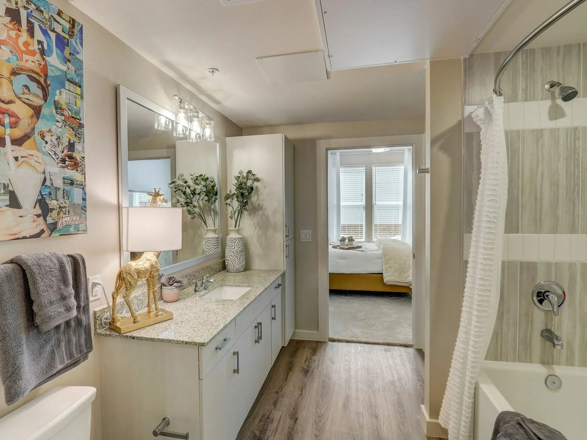 Bright bathroom vanity with granite counter, mirror, shower, and bedroom visible through open doorway