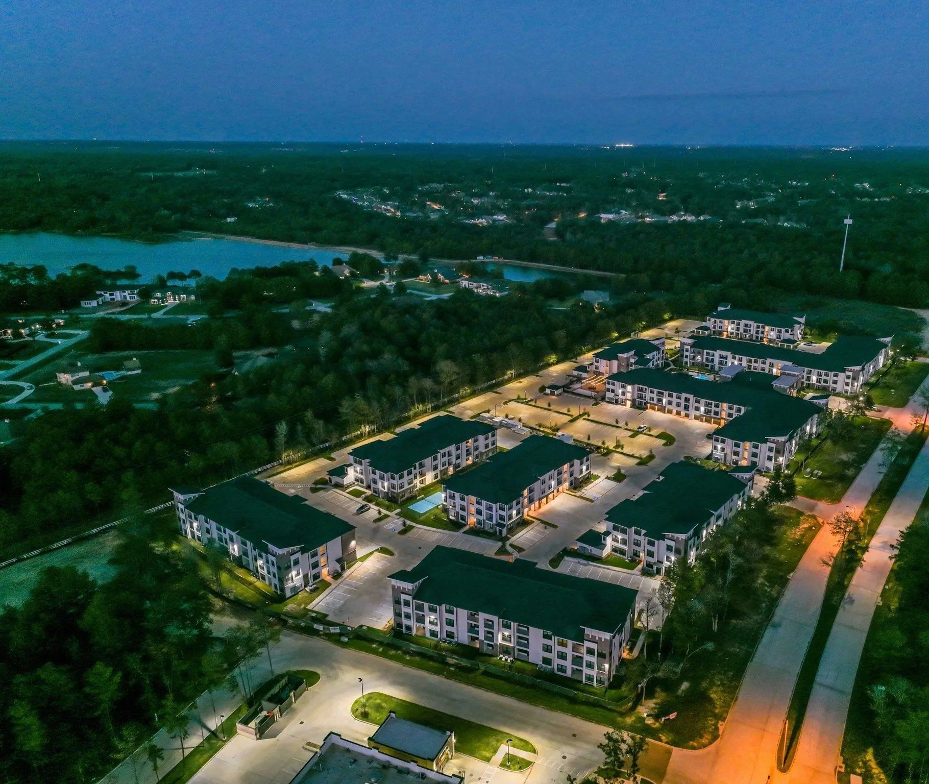 Aerial view of a lit apartment complex at dusk, surrounded by trees and roads.