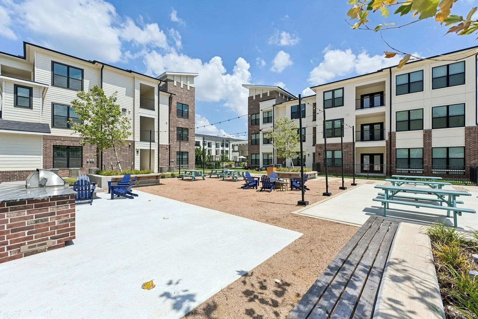 Modern apartment courtyard with blue chairs, picnic tables, brick paths, and sunny sky