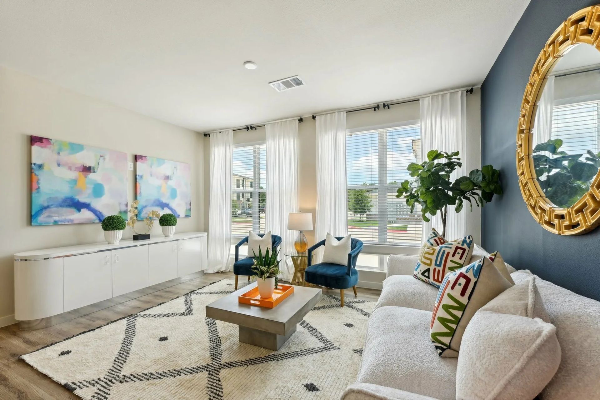Bright living room with blue chairs, white sofa, large windows, abstract art, and gold-framed mirror.
