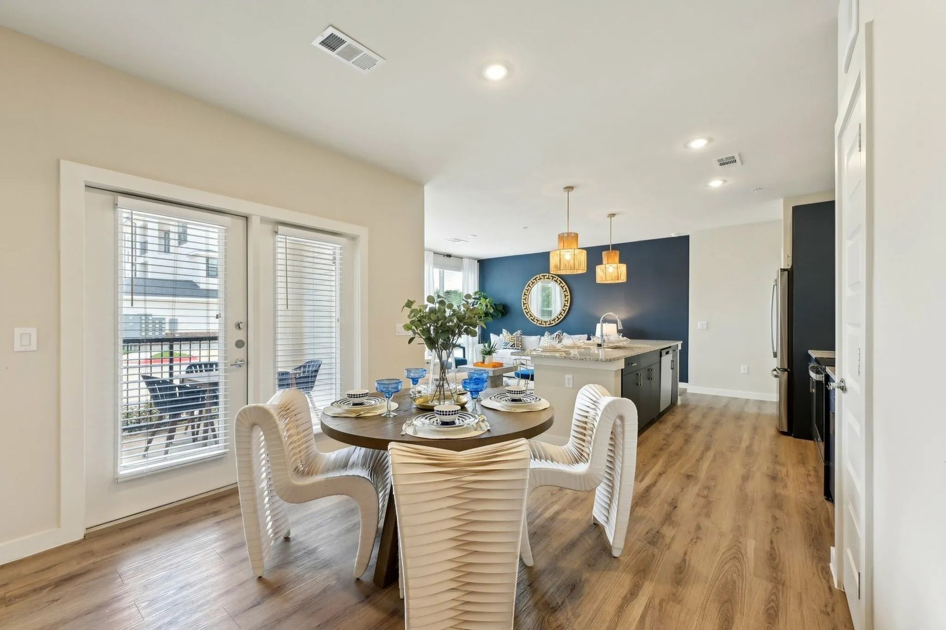 Bright dining area with round table, white chairs, blue accent wall, and open view to a kitchen/living space.