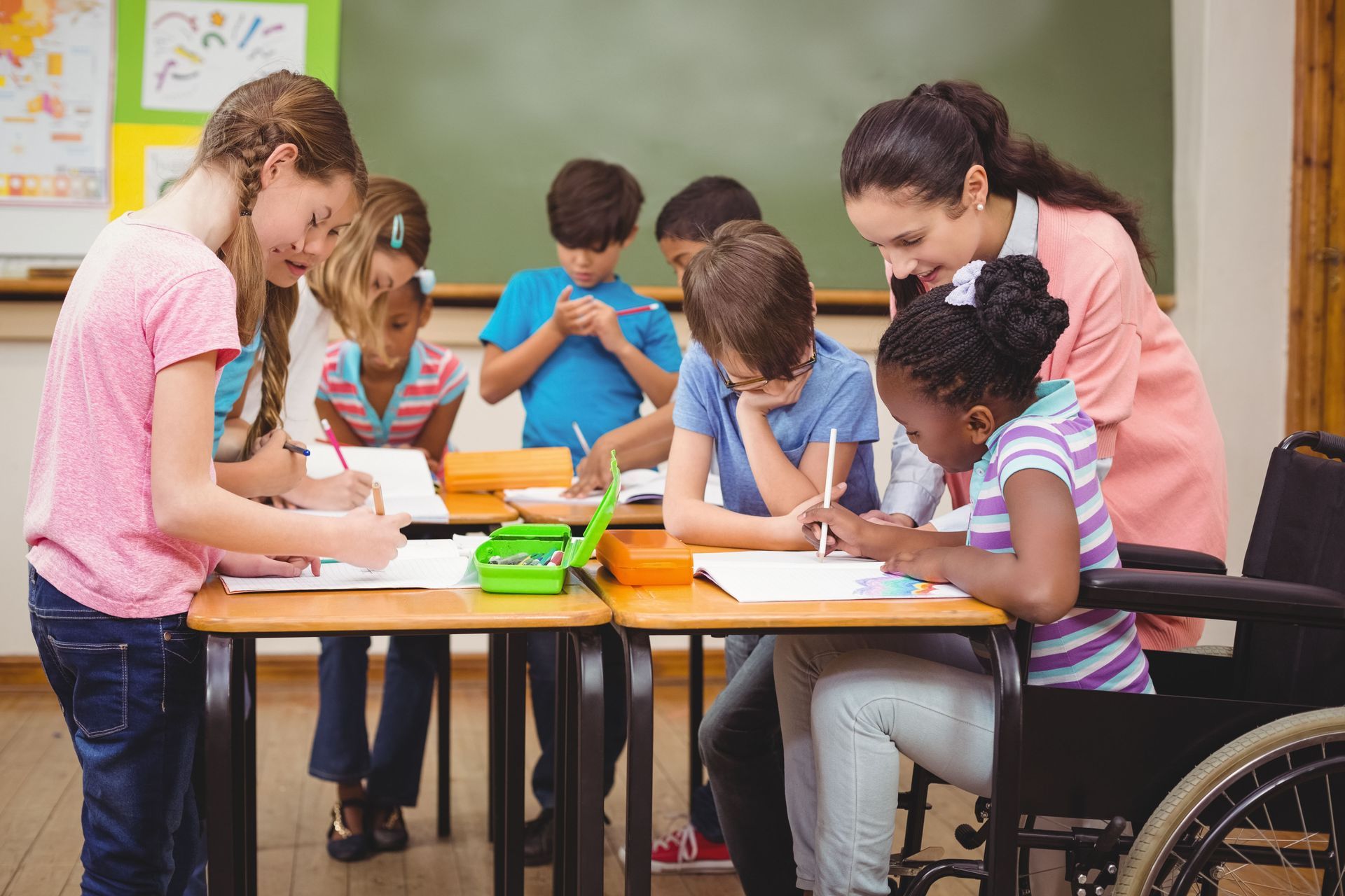 Teacher and students at desks, one student in a wheelchair, writing in a classroom.
