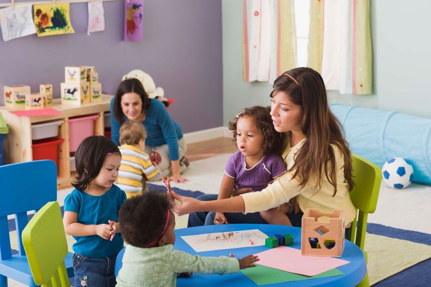 Children and teachers at a preschool: children drawing at a table while teachers observe.