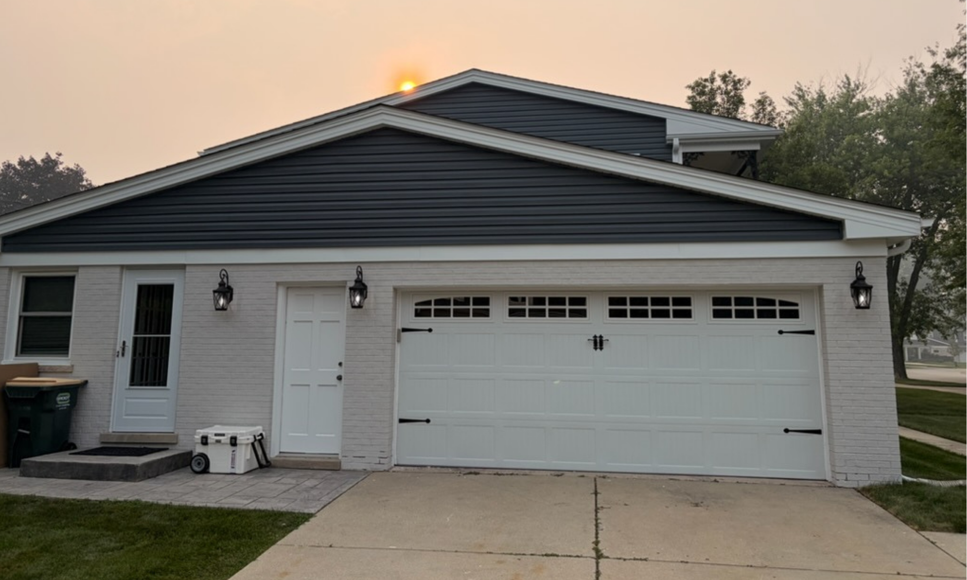 Single-story gray house with a white garage door and driveway at sunset