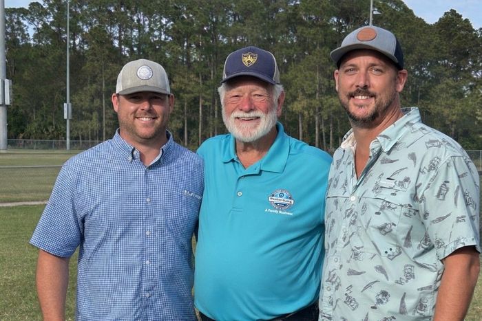 Three men smiling on a golf course, wearing caps and casual shirts, with trees in the background.