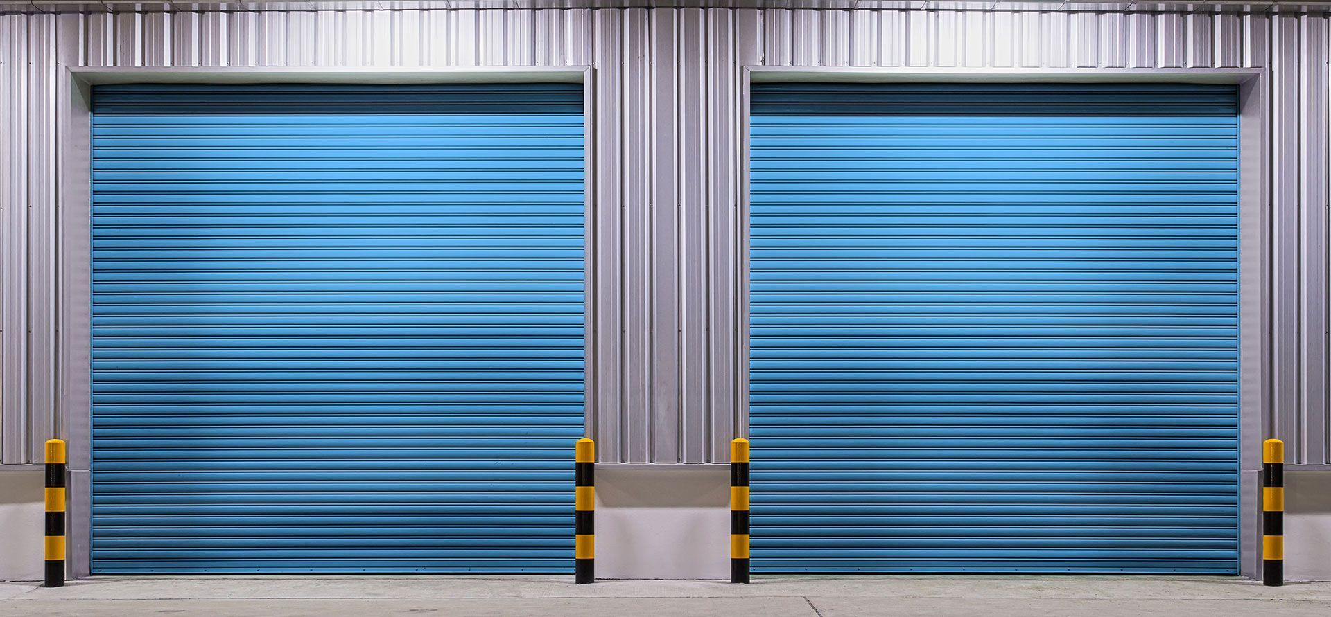 Two closed, bright blue industrial rolling garage doors on a metal warehouse wall with yellow and black safety bollards.