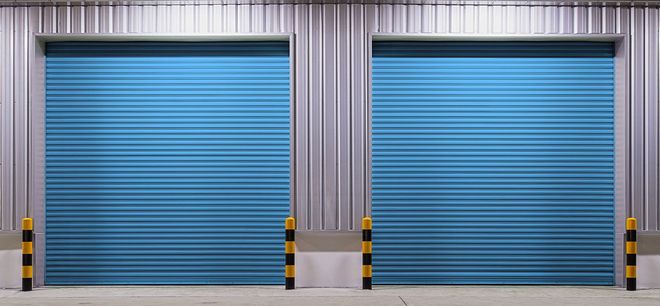 Two closed, bright blue industrial rolling garage doors on a metal warehouse wall with yellow and black safety bollards.